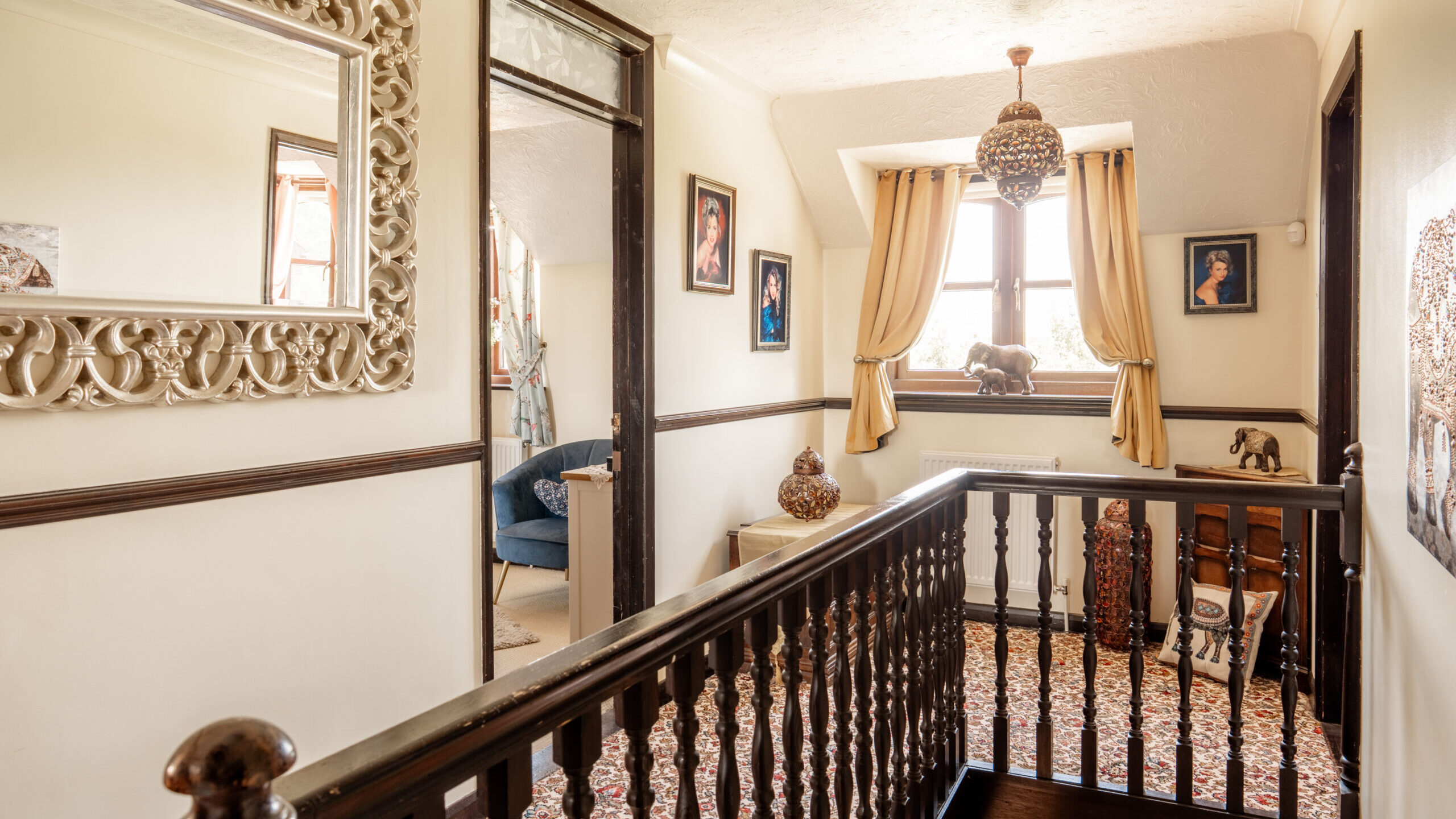 A bright hallway with patterned carpet, dark wood railing, framed art on cream walls, a large ornate mirror, decorative vases, and a window with gold curtains letting in natural light.