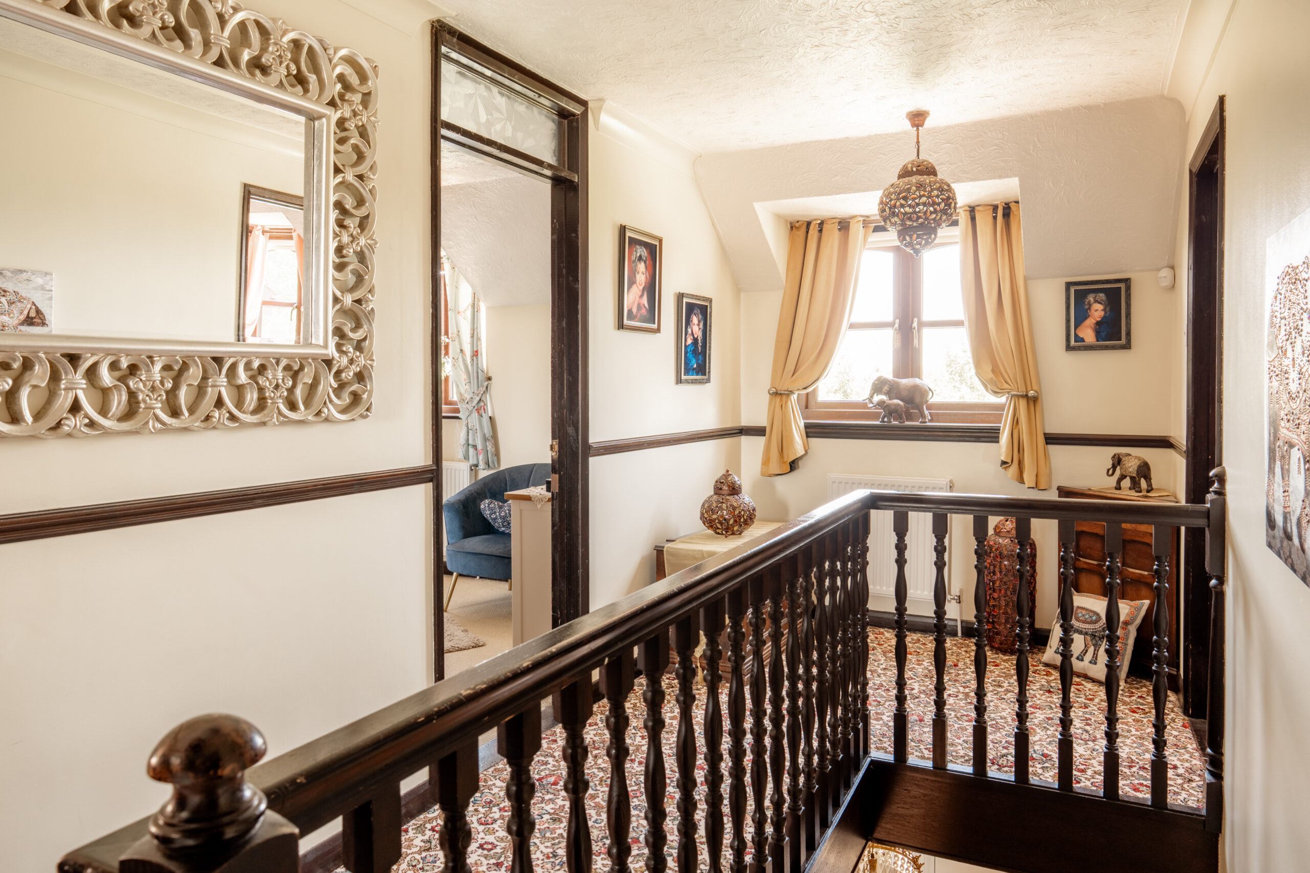 A bright hallway with patterned carpet, dark wood railing, framed art on cream walls, a large ornate mirror, decorative vases, and a window with gold curtains letting in natural light.