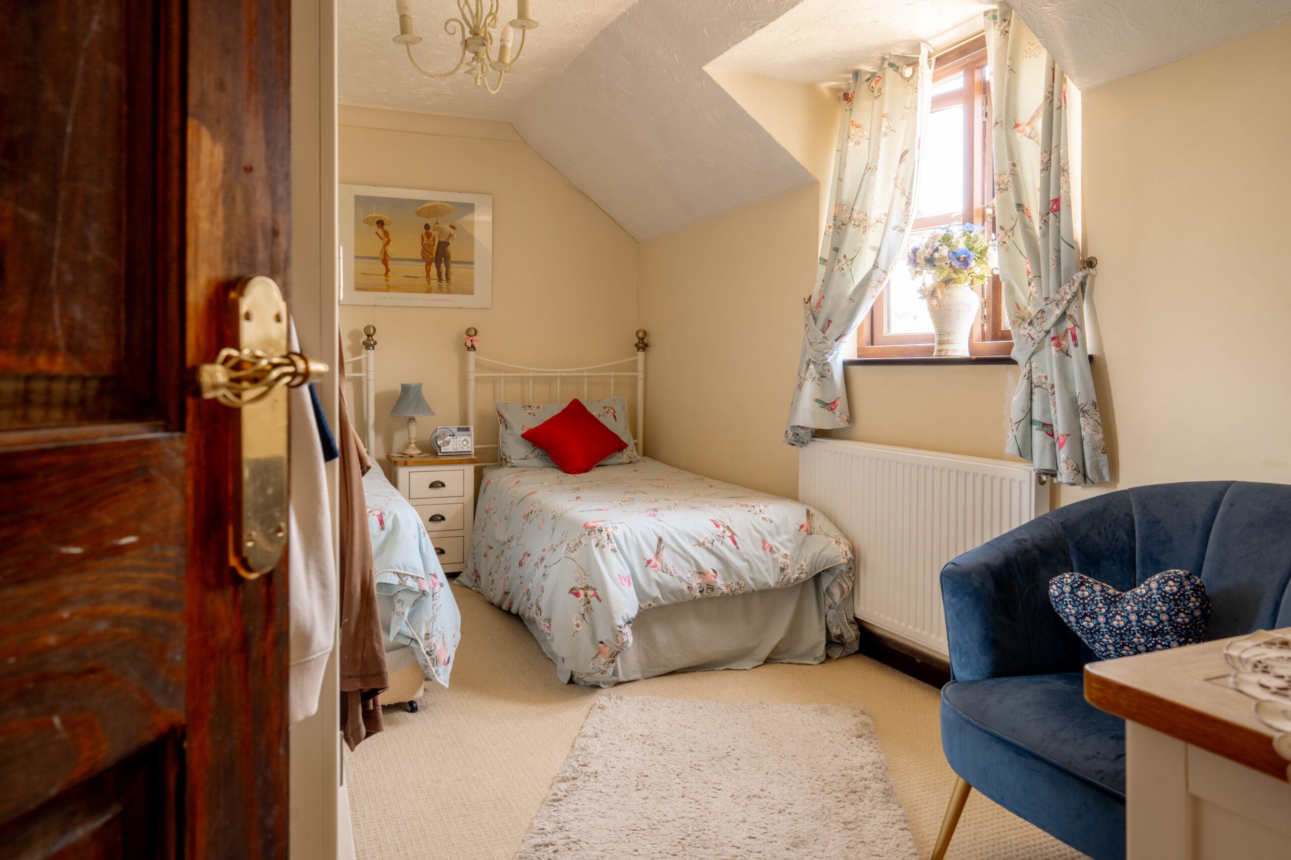 A cozy bedroom with a single bed, floral bedding, a red cushion, bedside tables, a window with floral curtains, a vase of flowers, blue armchair, and a rug on a light carpeted floor. Sunlight streams through the window.