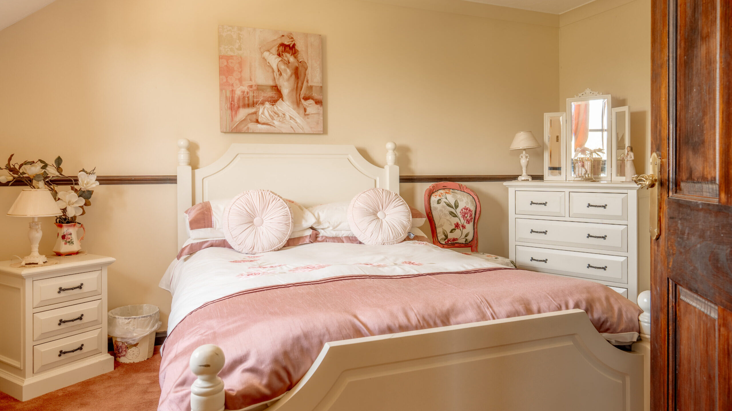 A cozy bedroom with a white bed, pink decorative pillows and bedding, matching nightstands and dresser, a floral chair, and soft lighting. A painting hangs above the bed, creating a warm and inviting atmosphere.