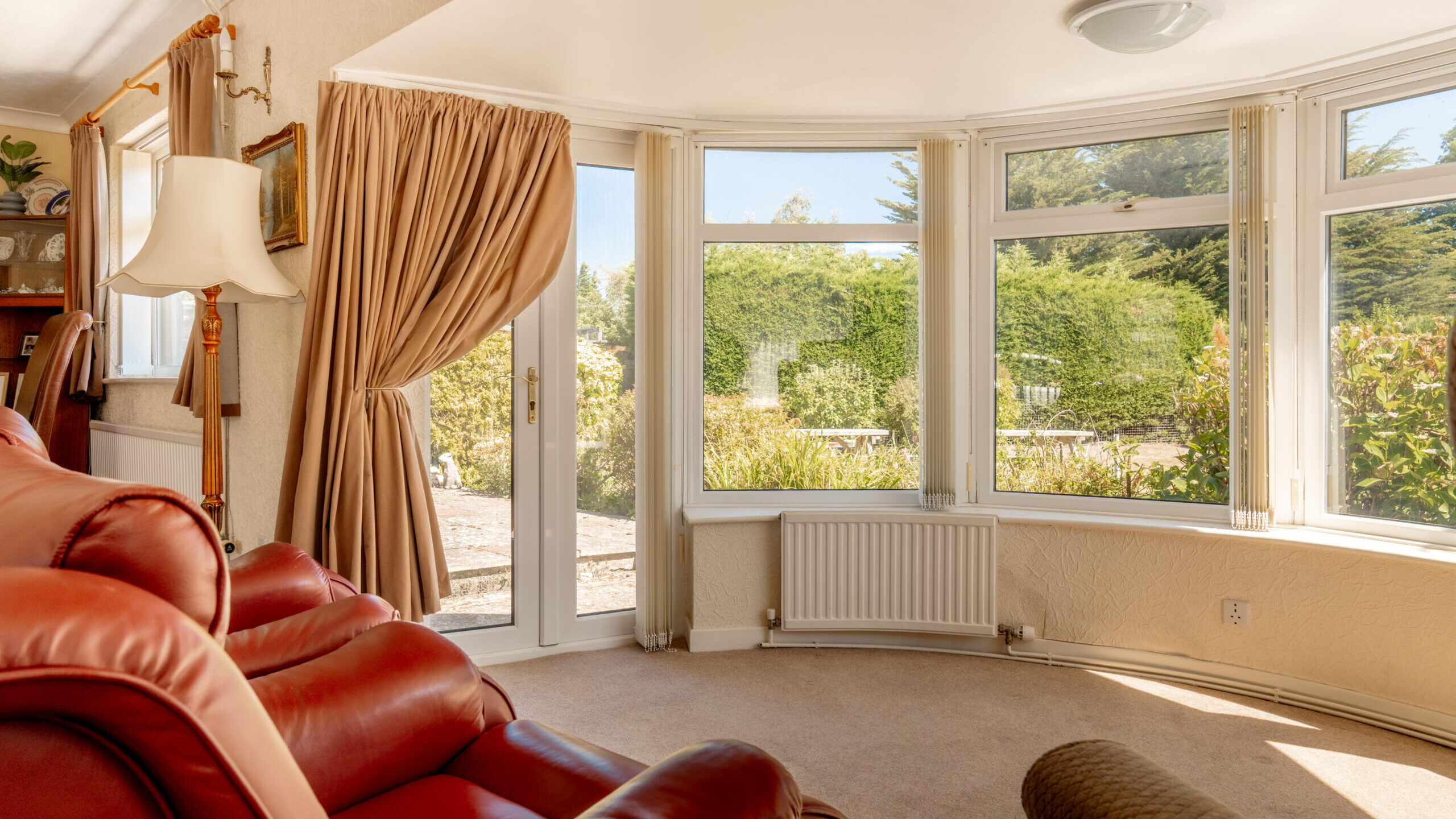 A cozy living room with red leather armchairs, beige carpet, large bay windows, and a glass door opening to a sunny garden with greenery outside. Curtains are drawn to one side, letting in natural light.