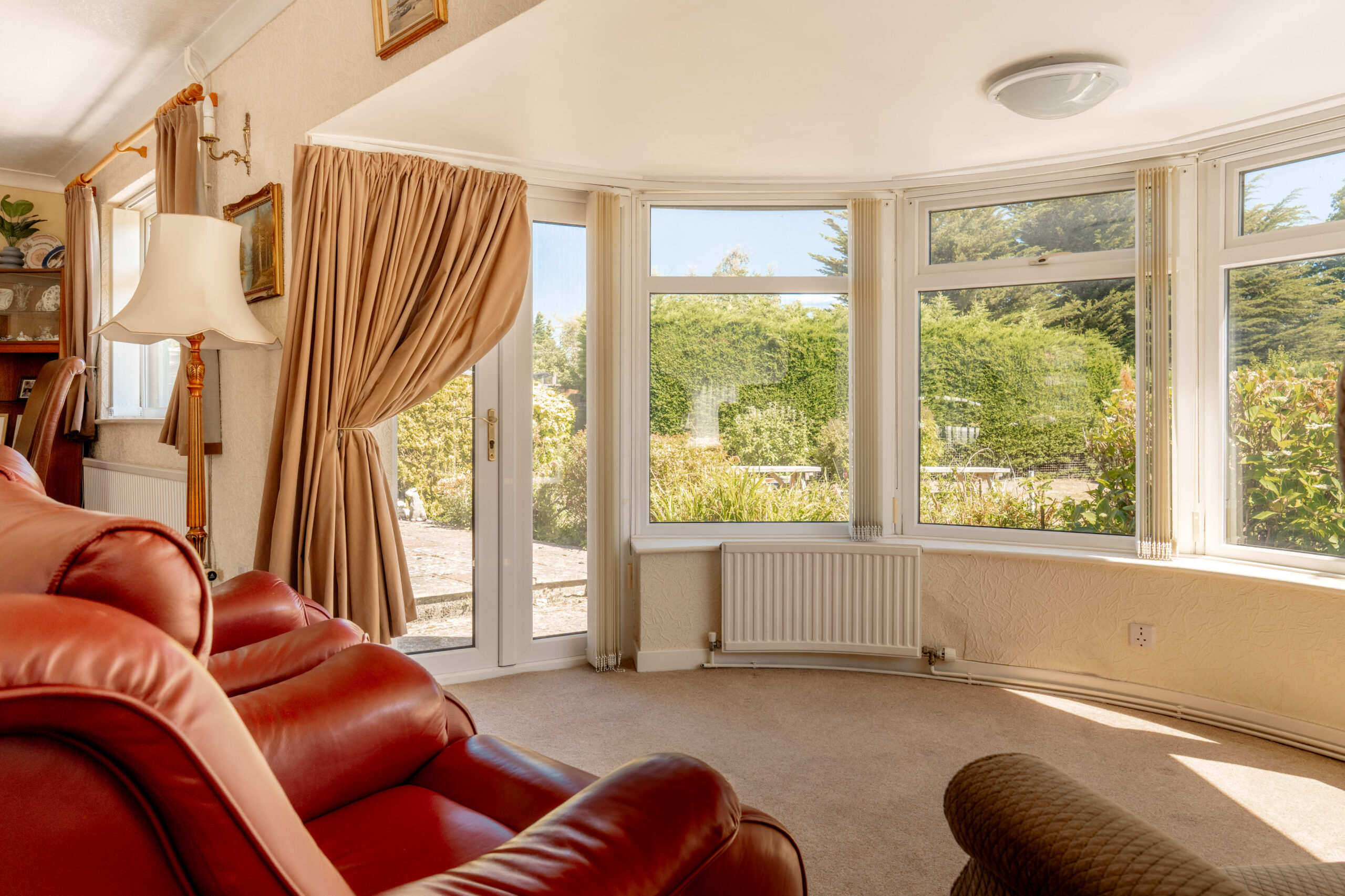 A cozy living room with red leather armchairs, beige carpet, large bay windows, and a glass door opening to a sunny garden with greenery outside. Curtains are drawn to one side, letting in natural light.