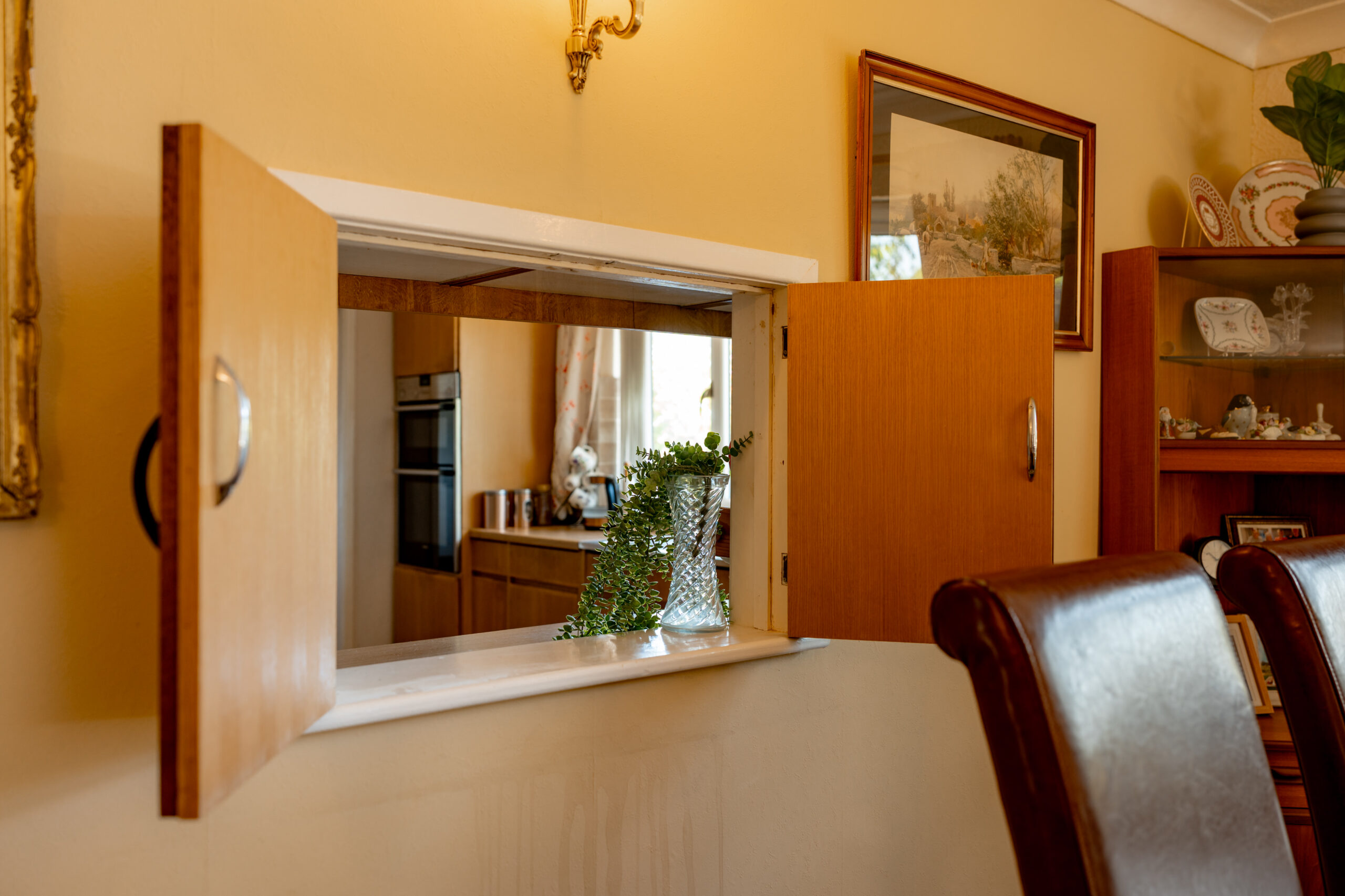 A view through an open wooden serving hatch between a kitchen and dining room, with a glass vase holding greenery on the sill and leather chairs in the foreground.