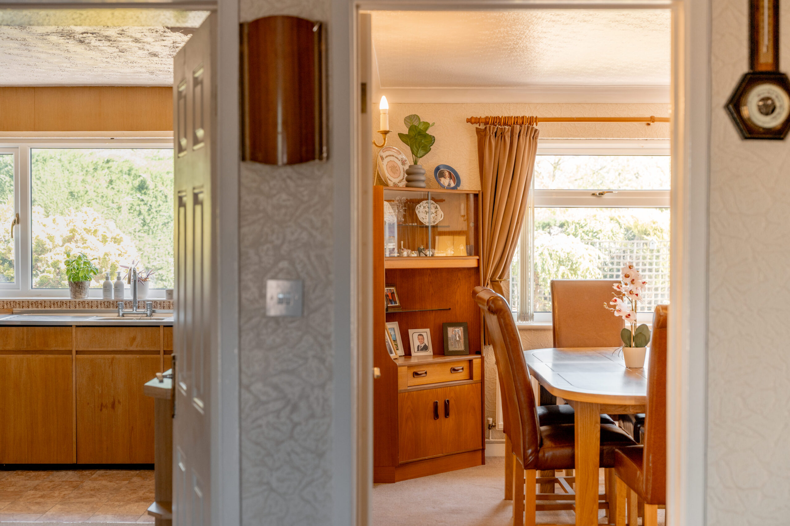 View through an open doorway into a dining room with a wooden table, chairs, and display cabinet; a bright kitchen with wooden cabinets and large windows is visible to the left.