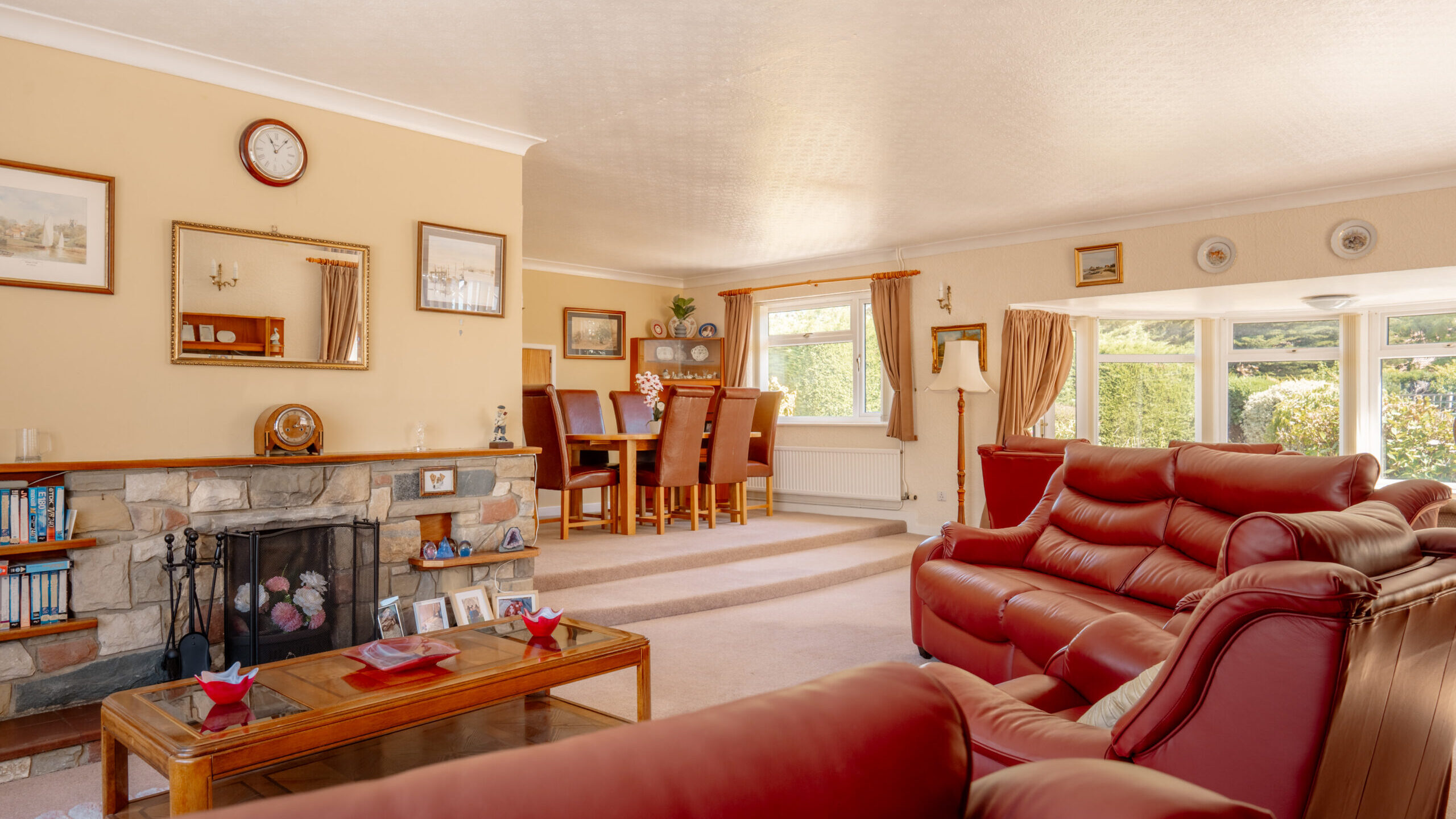 A spacious living room with red leather sofas, a stone fireplace, a wooden coffee table, and a dining area in the background. Large windows let in natural light, creating a bright and inviting atmosphere.