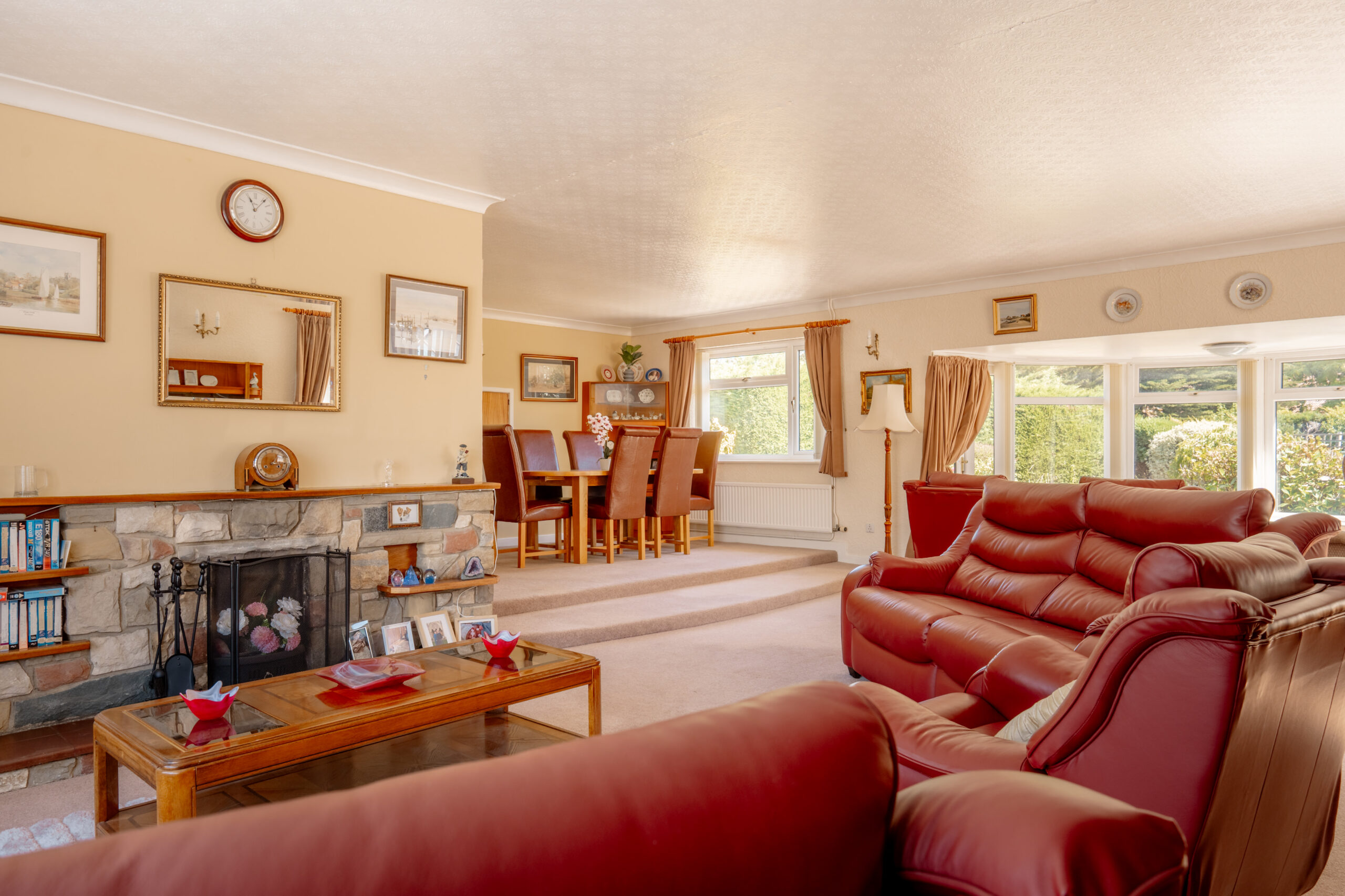 A spacious living room with red leather sofas, a stone fireplace, a wooden coffee table, and a dining area in the background. Large windows let in natural light, creating a bright and inviting atmosphere.
