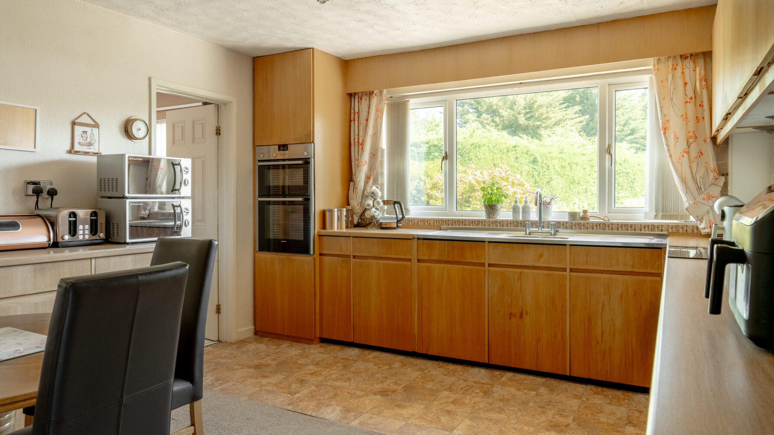 A spacious kitchen with wooden cabinets, a large window with floral curtains, built-in oven, microwave, toaster, and a dining table with black chairs, all brightly lit by natural light.