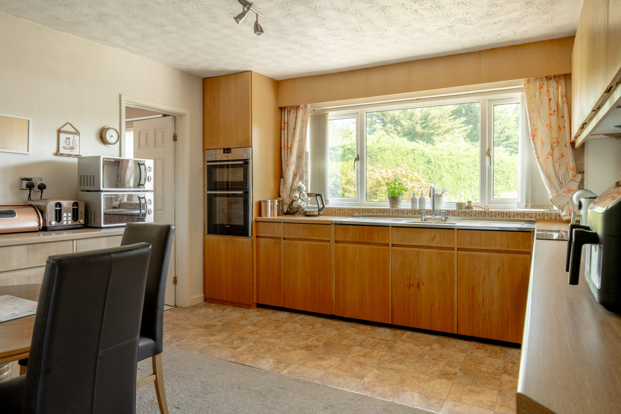 A spacious kitchen with wooden cabinets, a large window with floral curtains, built-in oven, microwave, toaster, and a dining table with black chairs, all brightly lit by natural light.