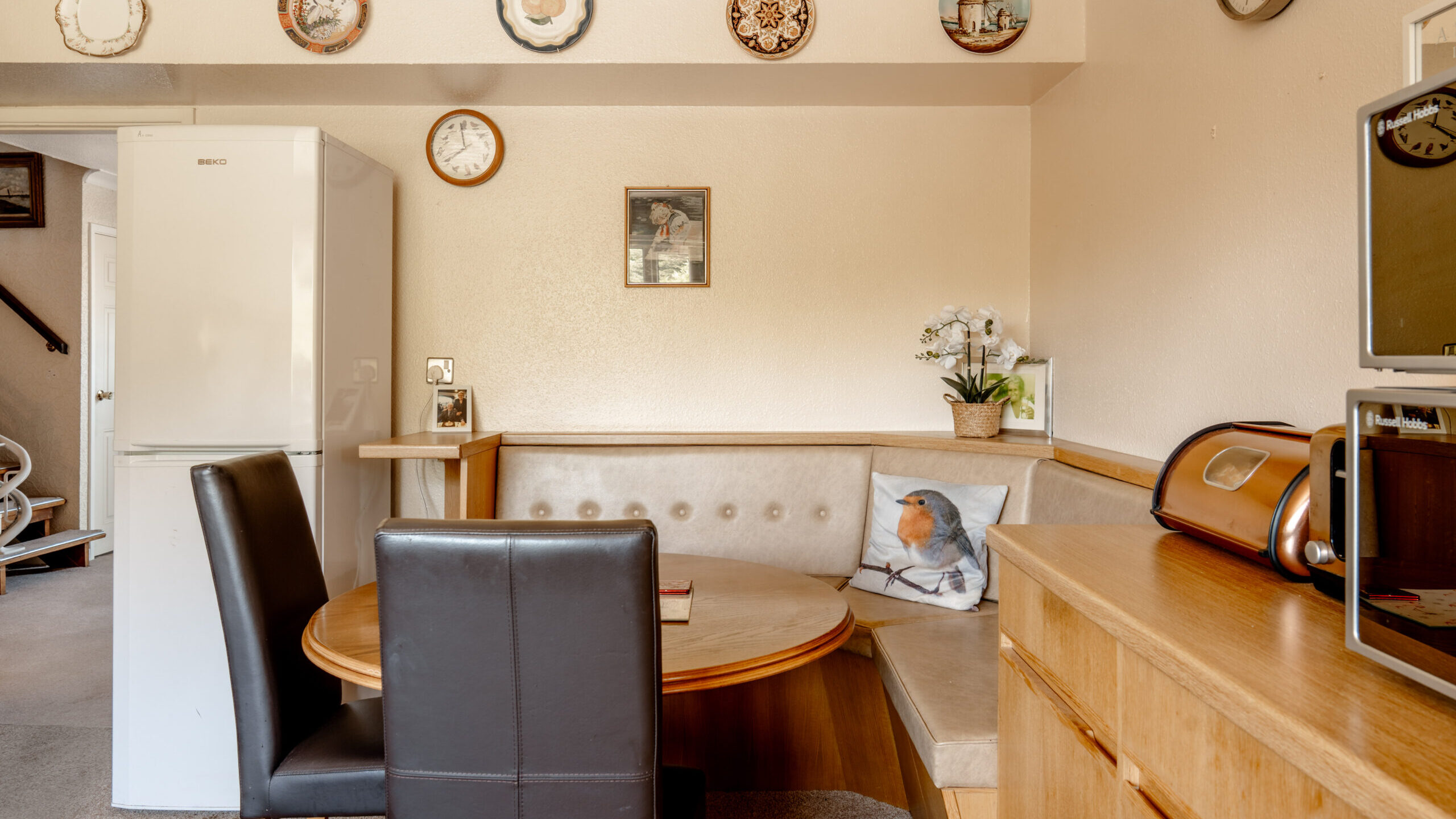 A cozy kitchen nook with a round wooden table, black and beige chairs, a corner bench with a bird cushion, a fridge, and a sideboard. Decorative plates and clocks hang on cream-colored walls.
