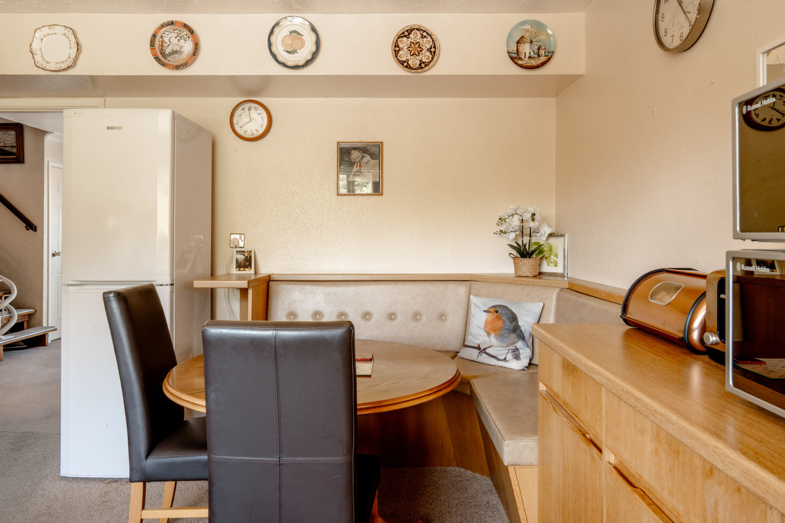 A cozy kitchen nook with a round wooden table, black and beige chairs, a corner bench with a bird cushion, a fridge, and a sideboard. Decorative plates and clocks hang on cream-colored walls.