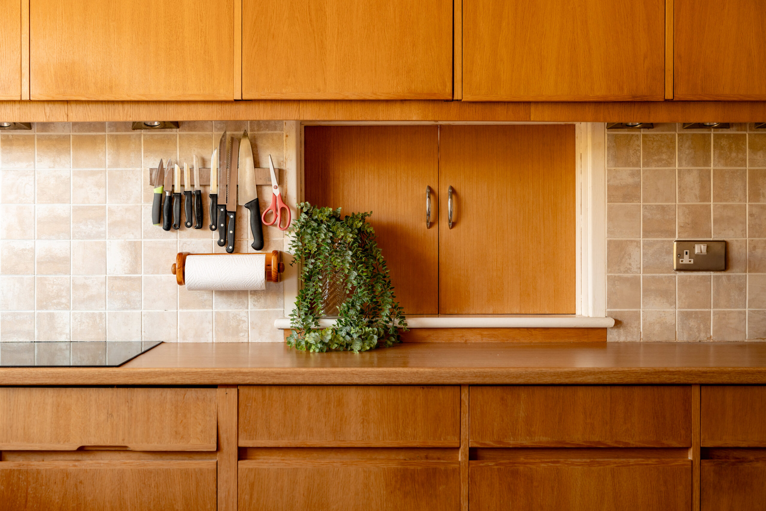 A tidy kitchen with light wood cabinets, beige tiled backsplash, knives and scissors hanging on a rack, paper towel holder, an electrical outlet, and a green leafy plant on the countertop.