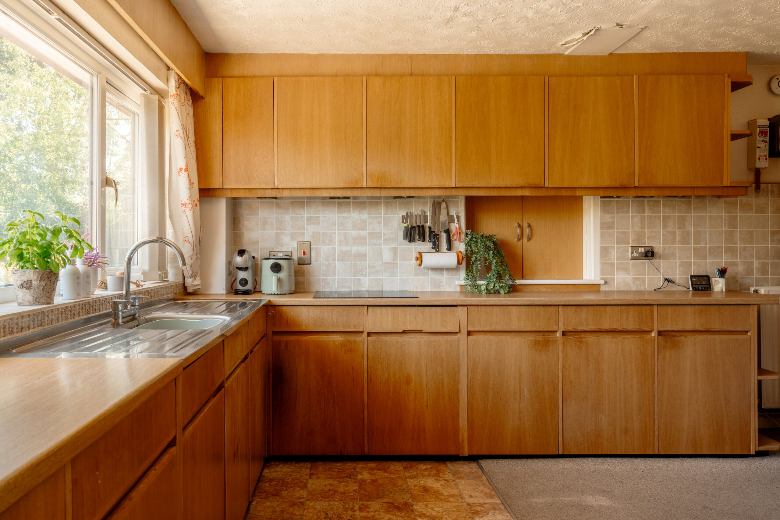 A spacious kitchen with wooden cabinets, a double sink under a window with potted plants, beige tiled backsplash, and various small appliances and utensils neatly arranged on the countertop.