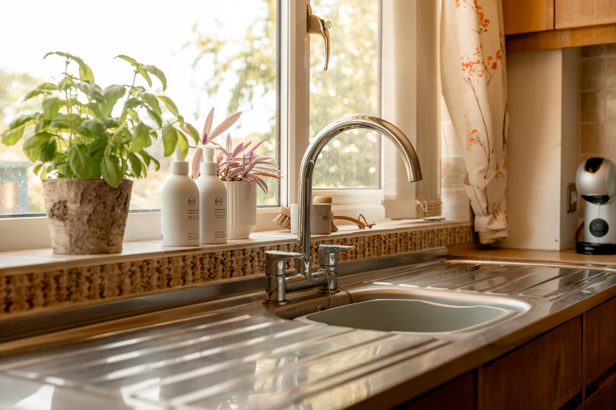 A sunlit kitchen sink with a silver faucet, potted plants, and soap dispensers on the windowsill. Light streams through a window framed by patterned curtains, illuminating the countertop.