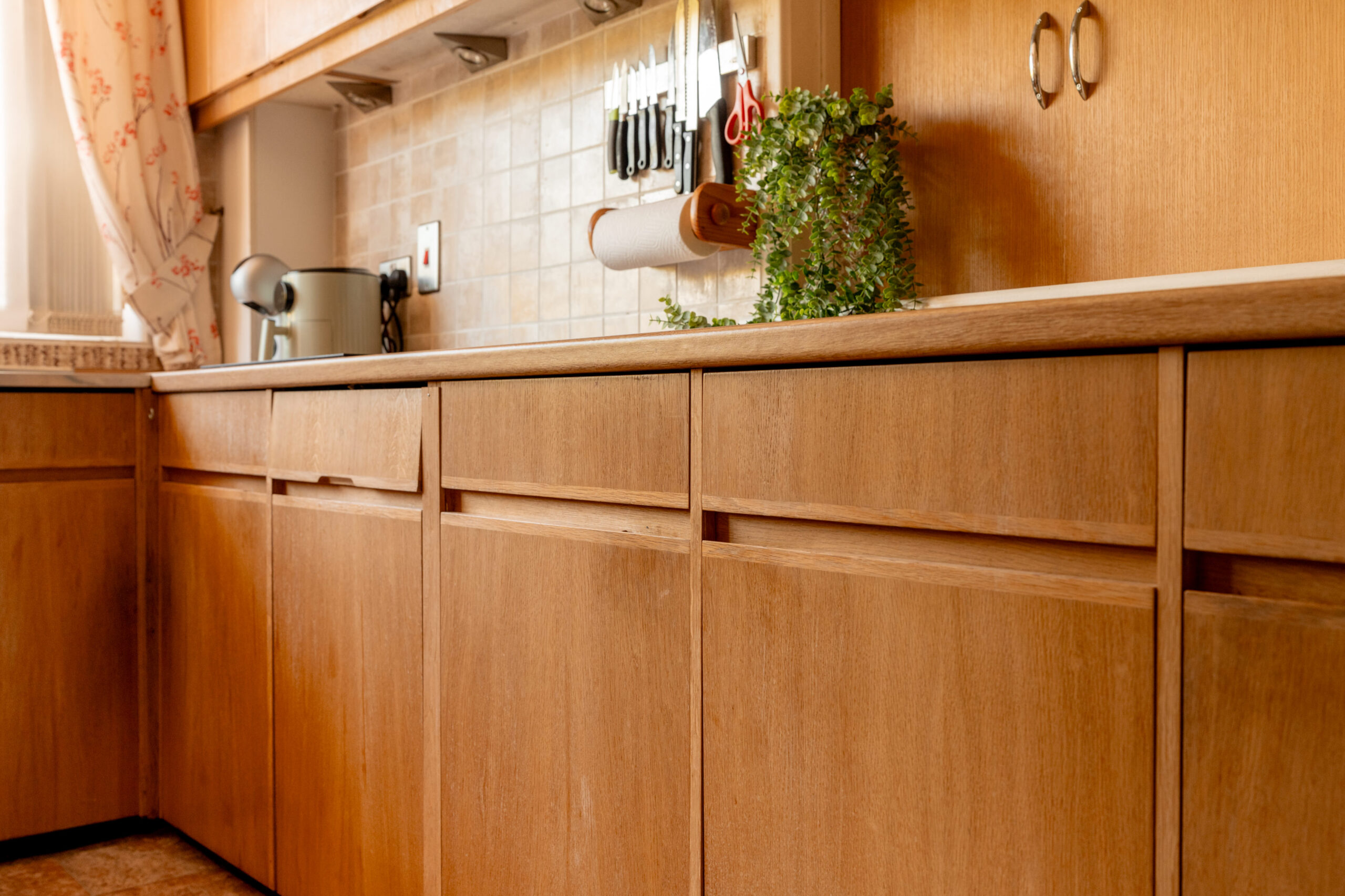 Close-up of wooden kitchen cabinets beneath a counter, with potted greenery, paper towel holder, utensils hanging on a rack, and light from a window with floral curtains illuminating the space.