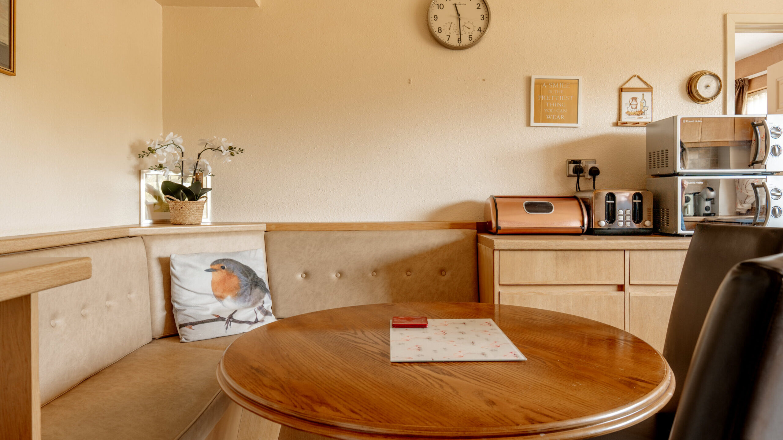 A cozy kitchen nook with a round wooden table, beige cushioned bench with a robin pillow, and a counter with a toaster, microwave, radio, and bread box. A wall clock and framed decor hang above.