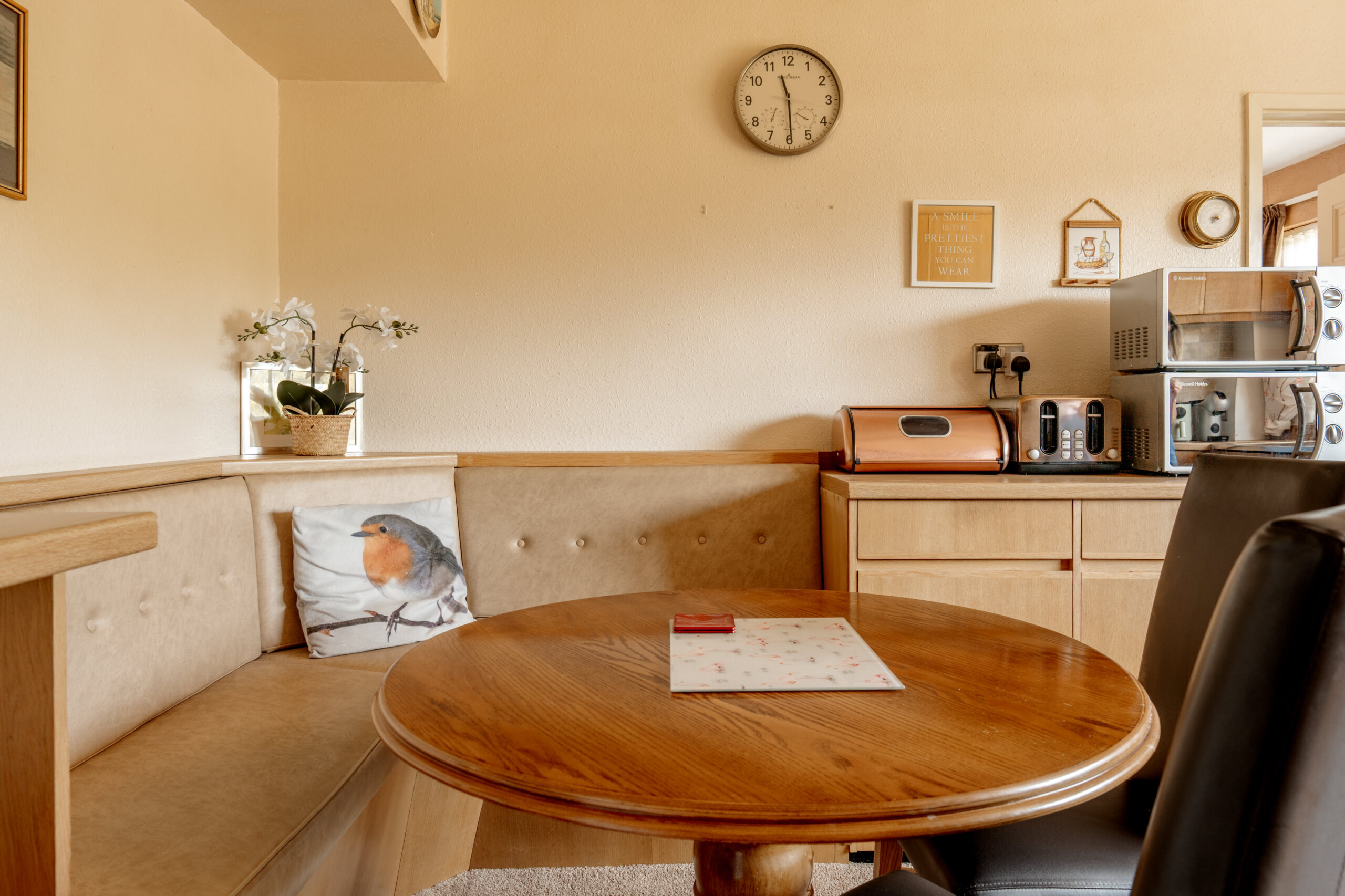 A cozy kitchen nook with a round wooden table, beige cushioned bench with a robin pillow, and a counter with a toaster, microwave, radio, and bread box. A wall clock and framed decor hang above.