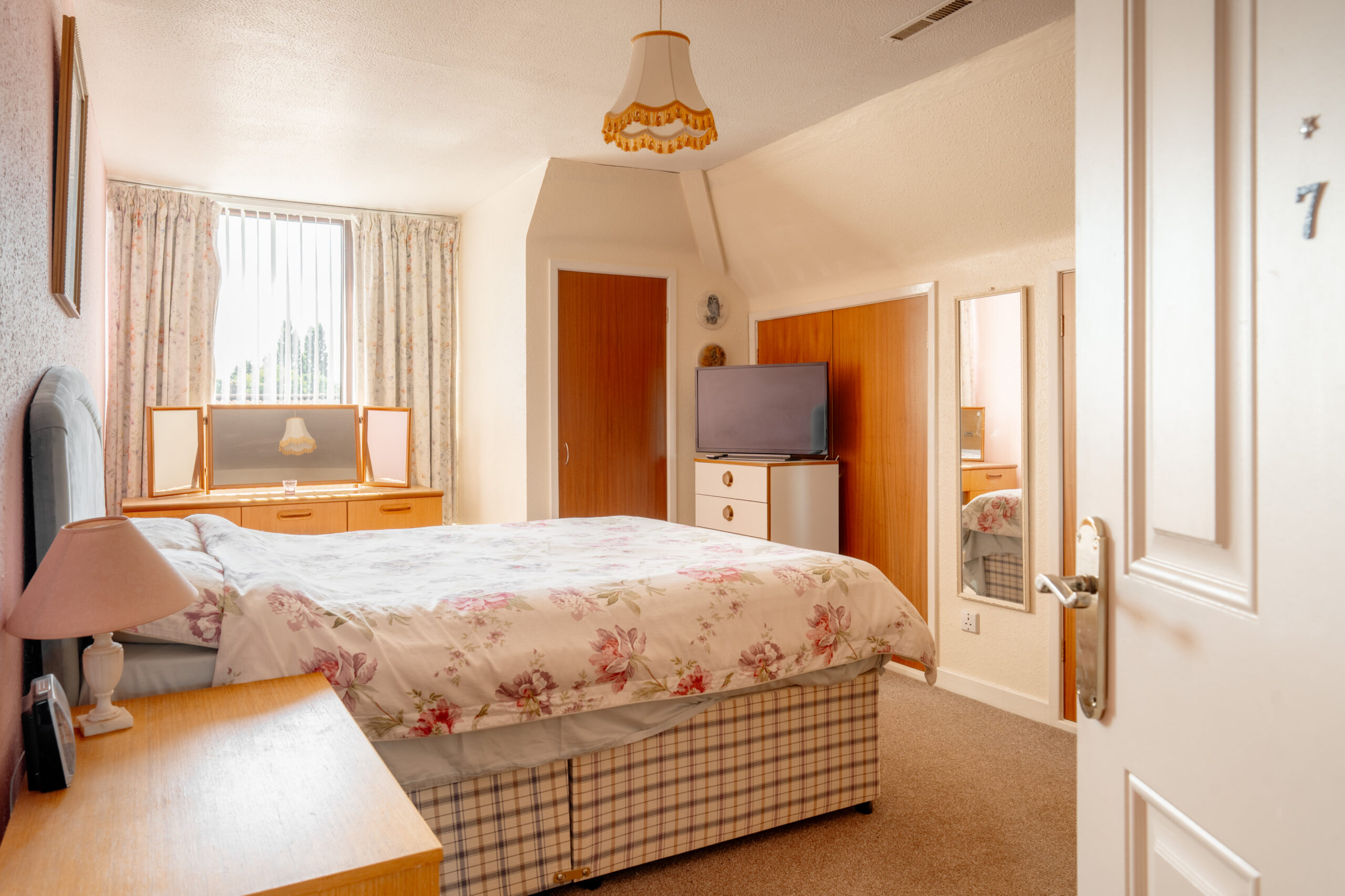 A cozy bedroom with a double bed covered in floral bedding, wooden furniture, a dressing table by the window, a TV on a chest of drawers, a mirror, and soft lighting from a ceiling lamp. The door is partially open.