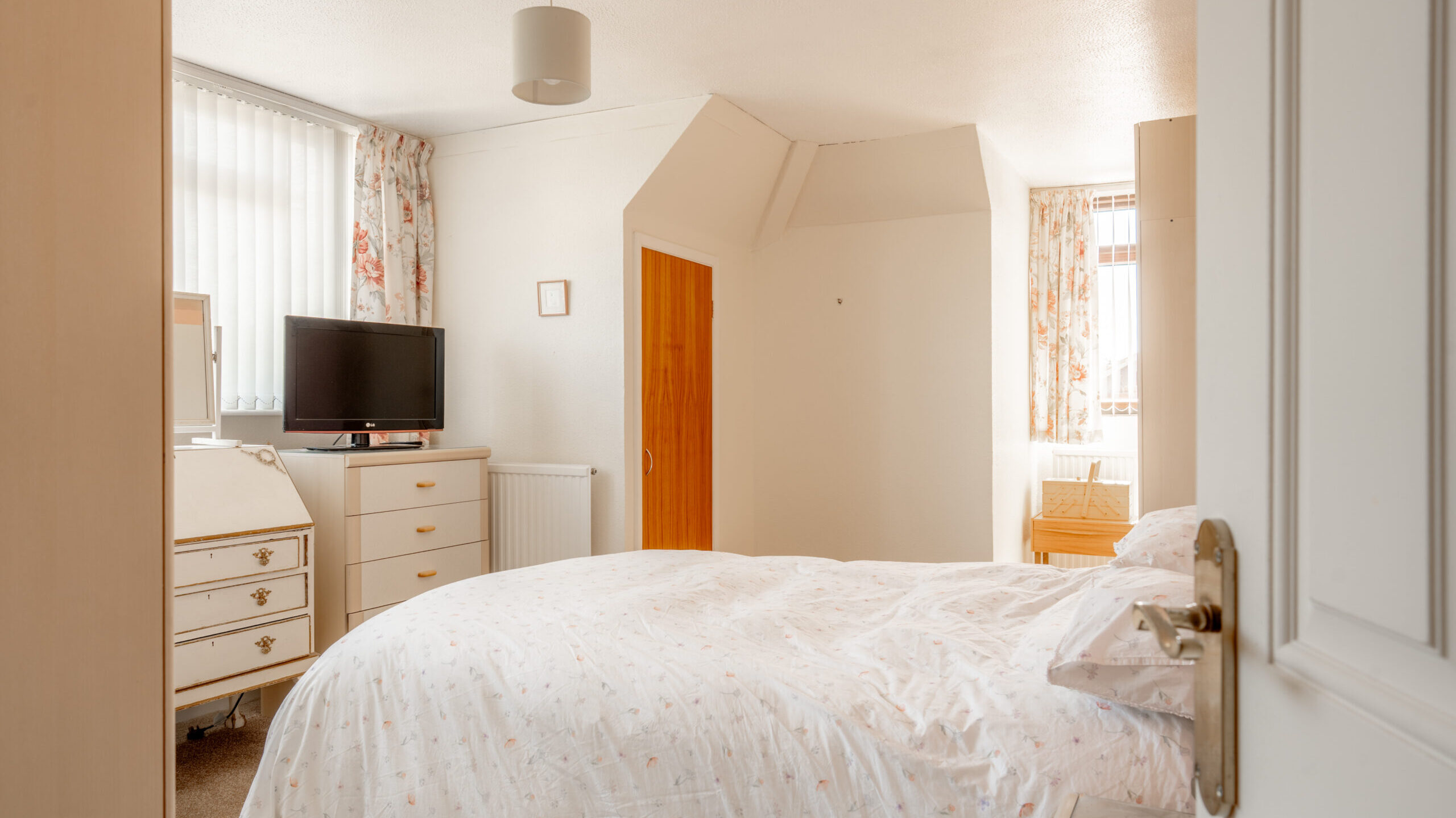 A bright bedroom with a double bed, floral curtains, a TV on a dresser, a wooden door, and a vanity. Light streams in from two windows, creating a warm and inviting atmosphere.
