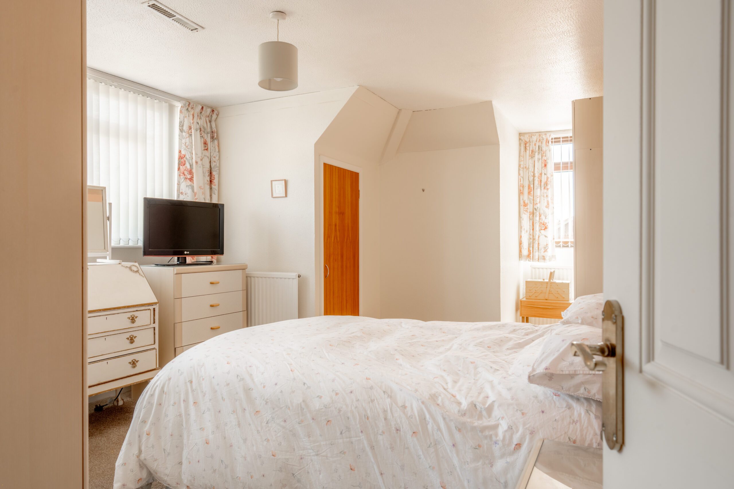 A bright bedroom with a double bed, floral curtains, a TV on a dresser, a wooden door, and a vanity. Light streams in from two windows, creating a warm and inviting atmosphere.
