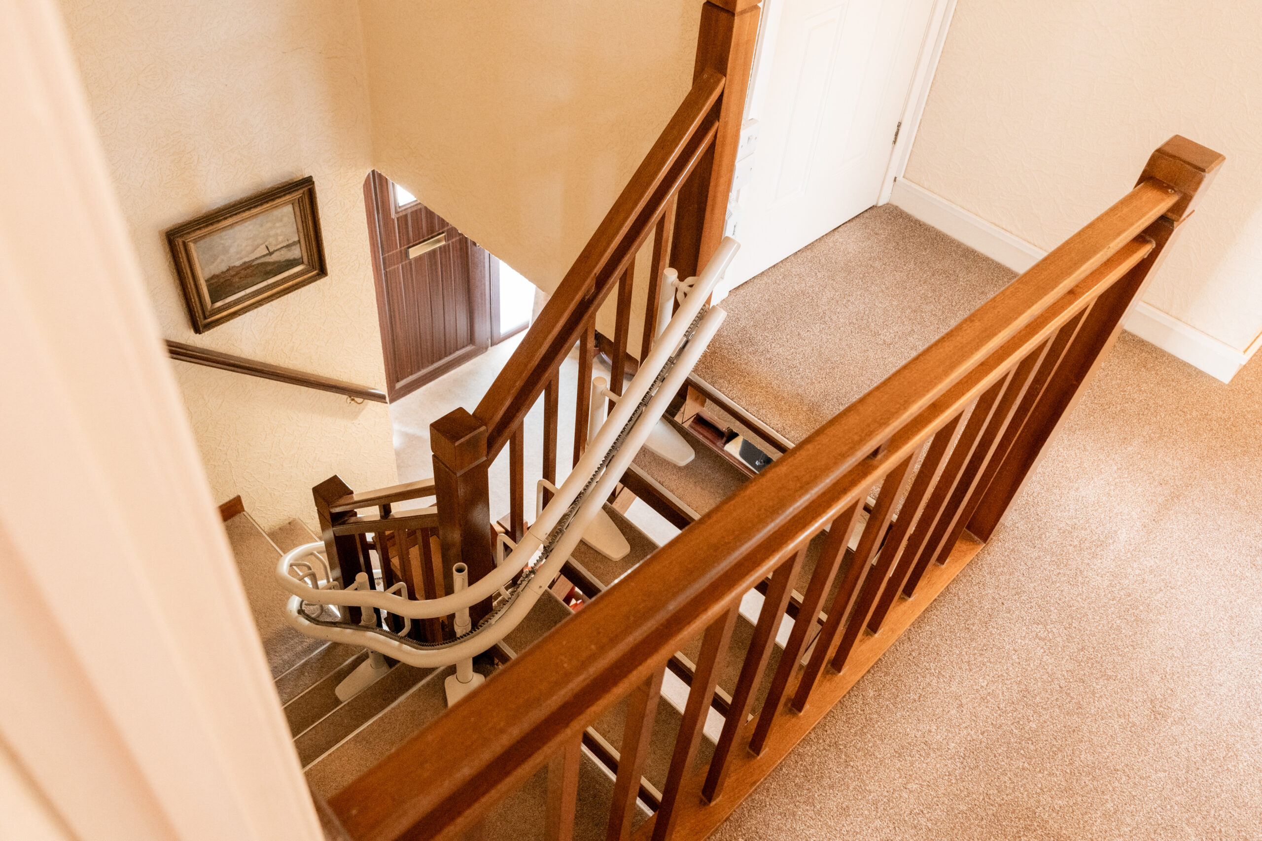 A stairlift is installed along a carpeted staircase with wooden railings in a home, leading down to a hallway with a framed picture on the wall and a wooden front door.
