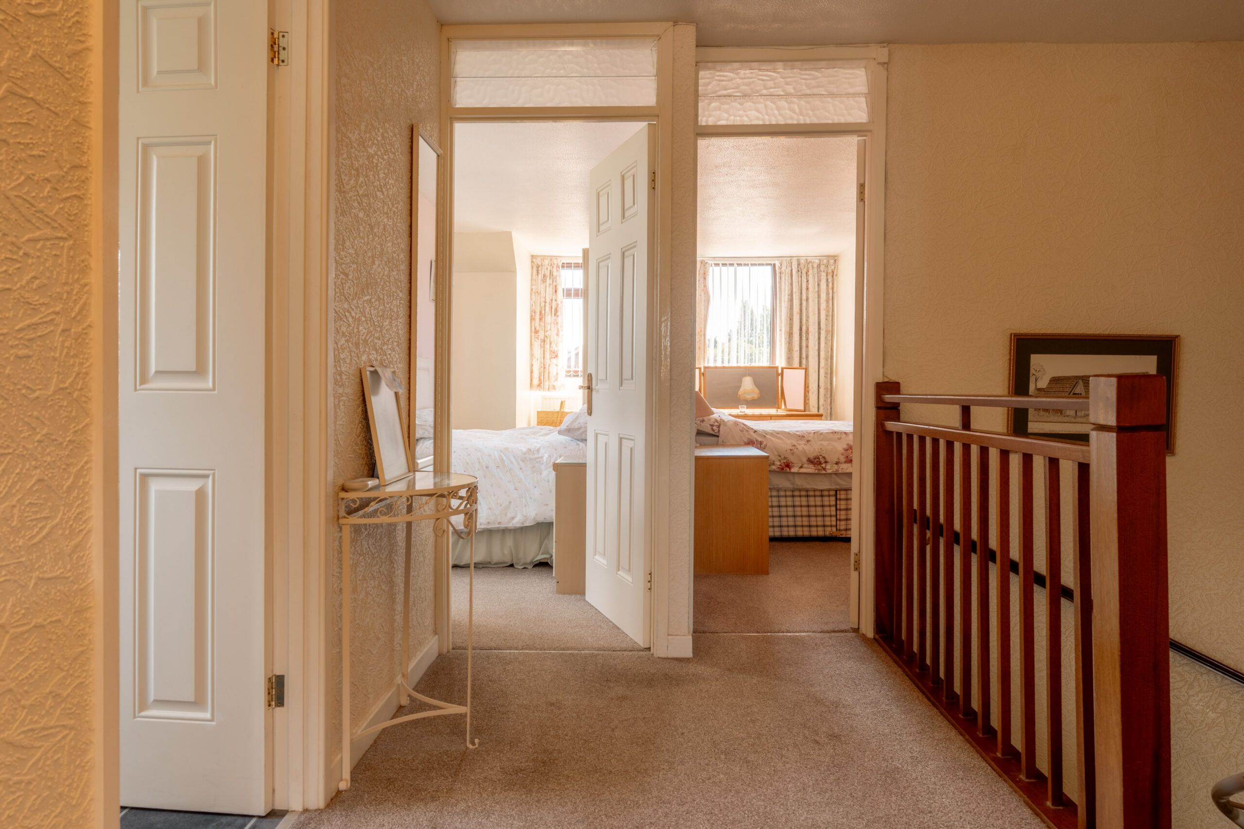 A carpeted hallway with a wooden railing leads to two open doors, each revealing bright bedrooms with beds, light-colored walls, and windows letting in natural light. A small table with a mirror sits on the left.
