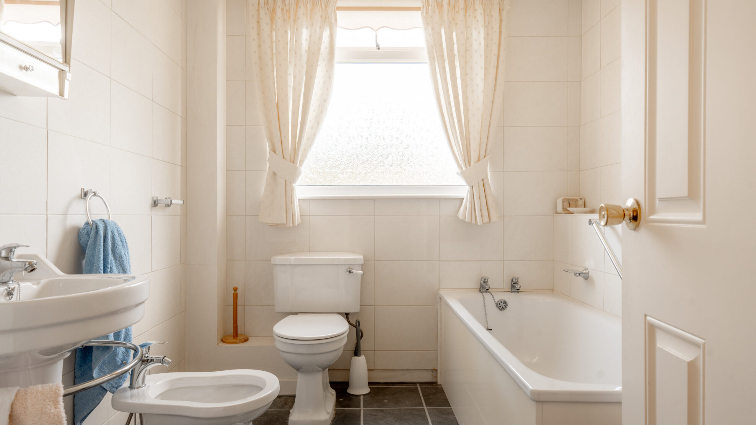 Bright, clean bathroom with a toilet, bidet, sink, and bathtub. Cream-colored tiles, white fixtures, and a large window with sheer curtains let in natural light. Blue towel, soap, and toilet brush visible.