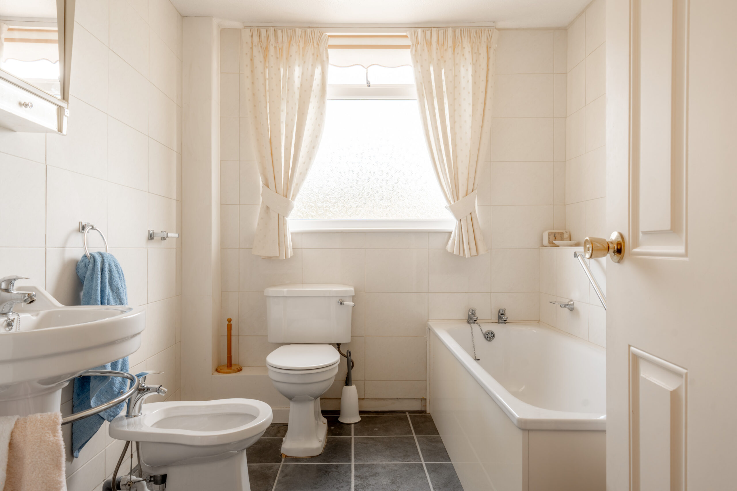 Bright, clean bathroom with a toilet, bidet, sink, and bathtub. Cream-colored tiles, white fixtures, and a large window with sheer curtains let in natural light. Blue towel, soap, and toilet brush visible.