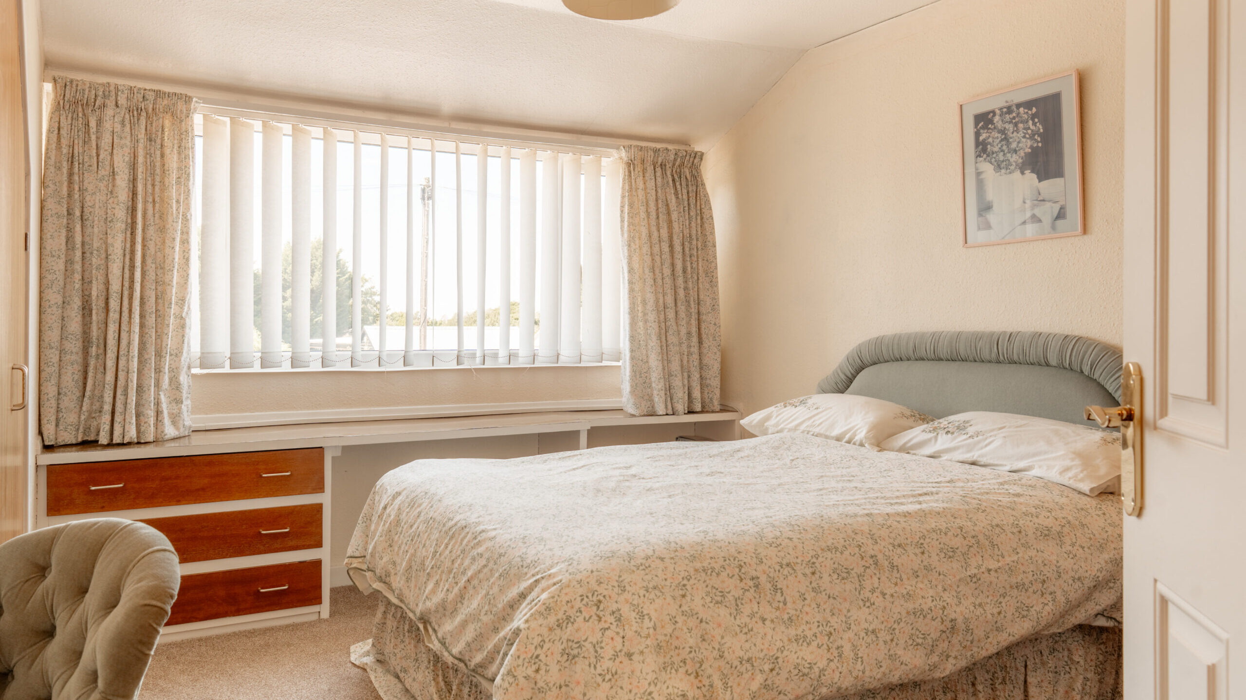 A cozy bedroom with a double bed, floral bedding, a tufted chair, a large window with vertical blinds and patterned curtains, built-in drawers, and framed artwork on a cream-colored wall.