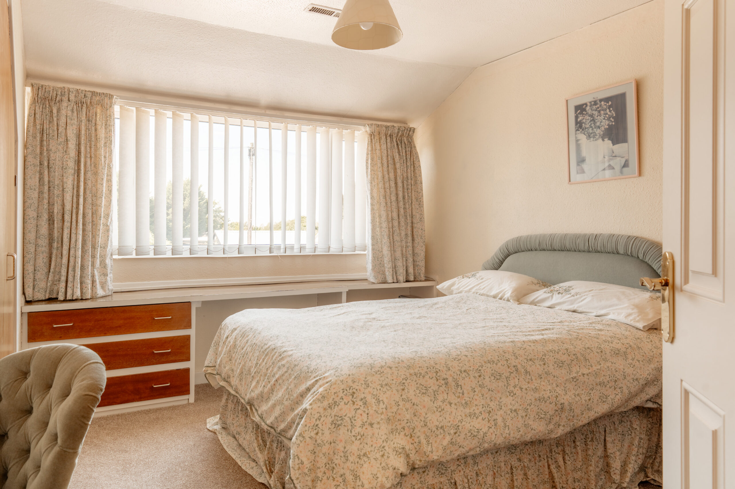 A cozy bedroom with a double bed, floral bedding, a tufted chair, a large window with vertical blinds and patterned curtains, built-in drawers, and framed artwork on a cream-colored wall.