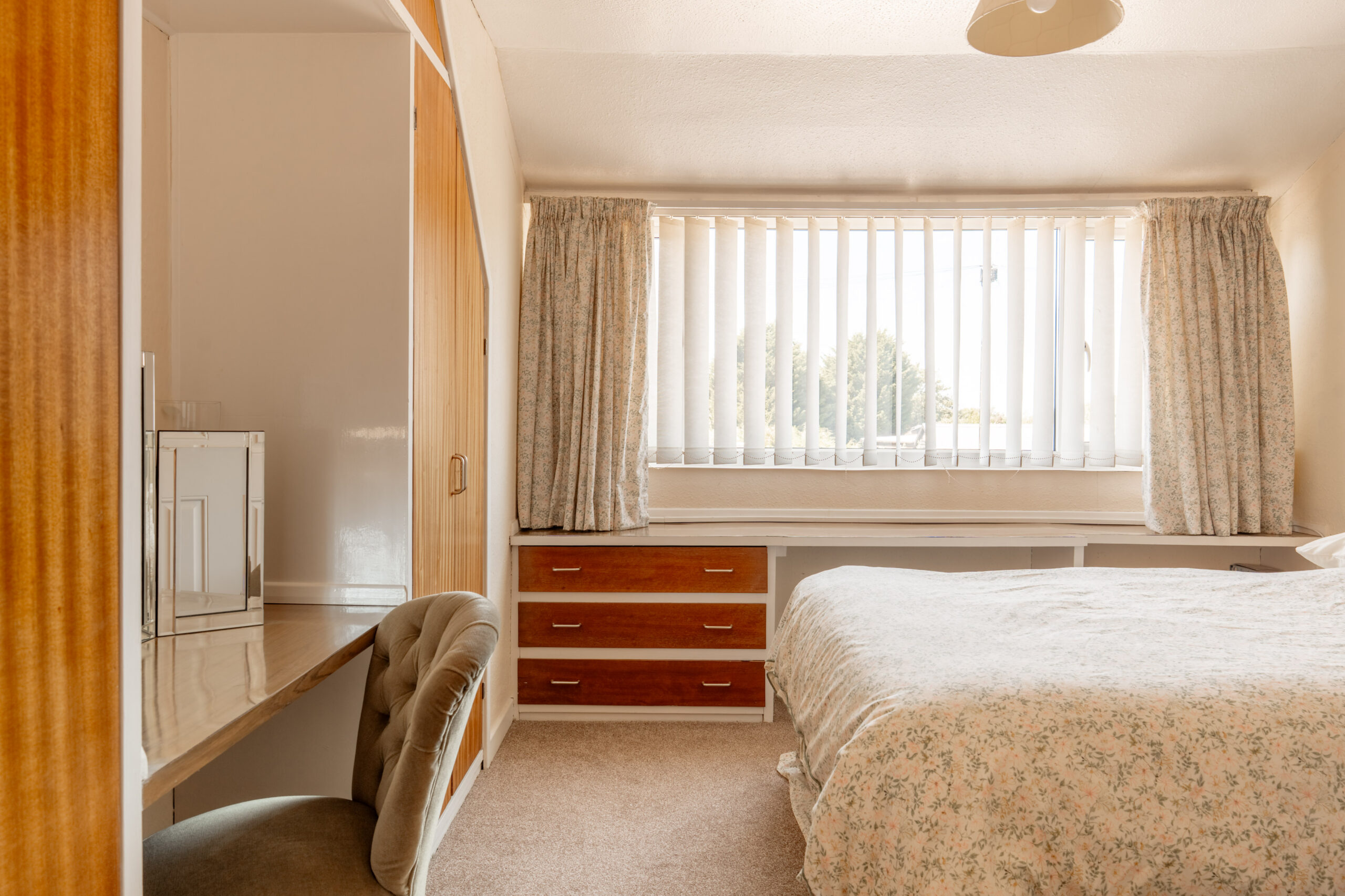 A cozy bedroom with a large window covered by vertical blinds, floral curtains, a bed with a patterned duvet, built-in wooden furniture, and a desk with a padded chair on beige carpet.