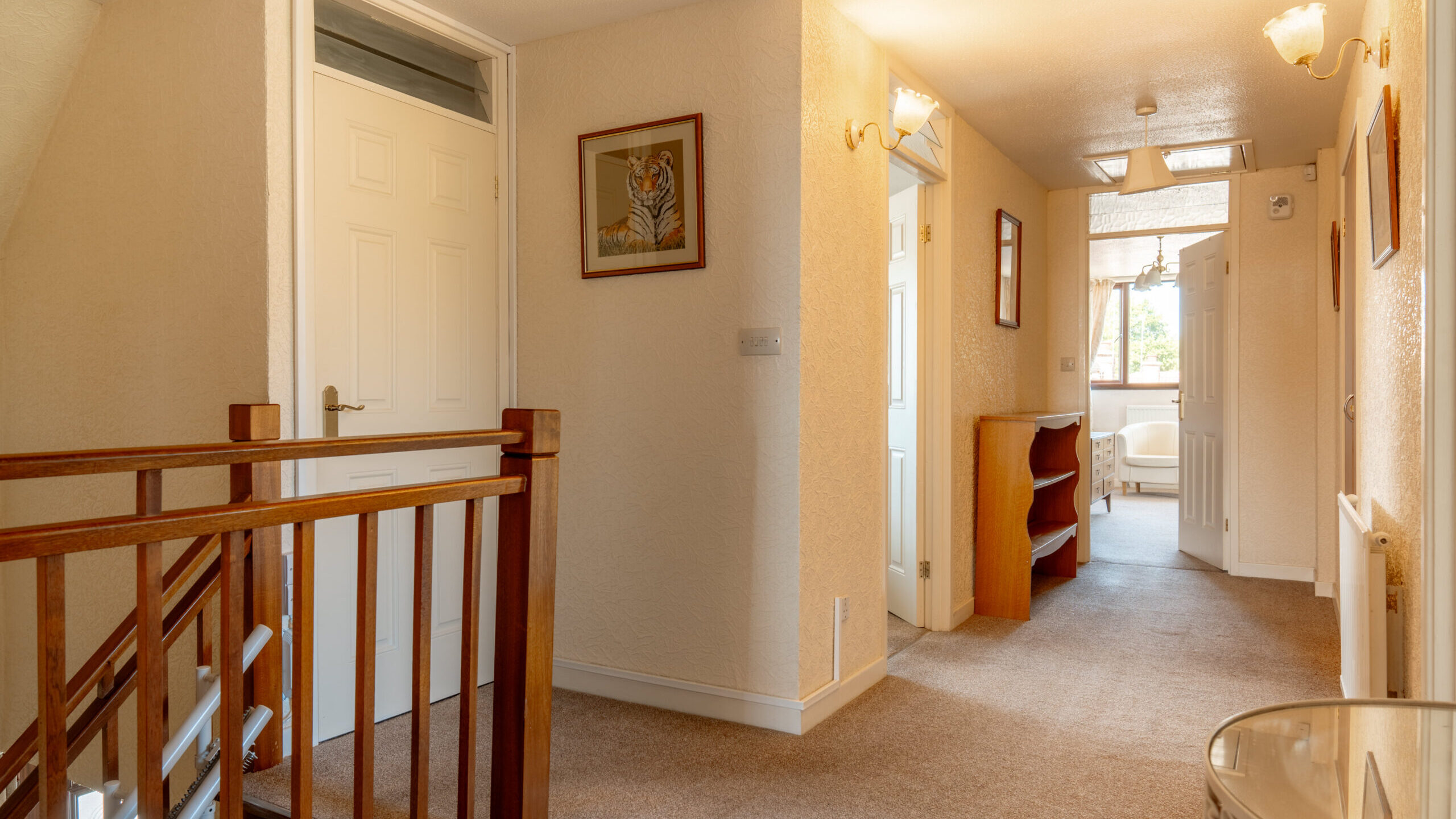 A well-lit hallway in a house with beige walls, carpeted floor, wooden stair railing, several doors, a framed tiger picture on the wall, and a small glass table in the corner. Light streams in from a distant window.
