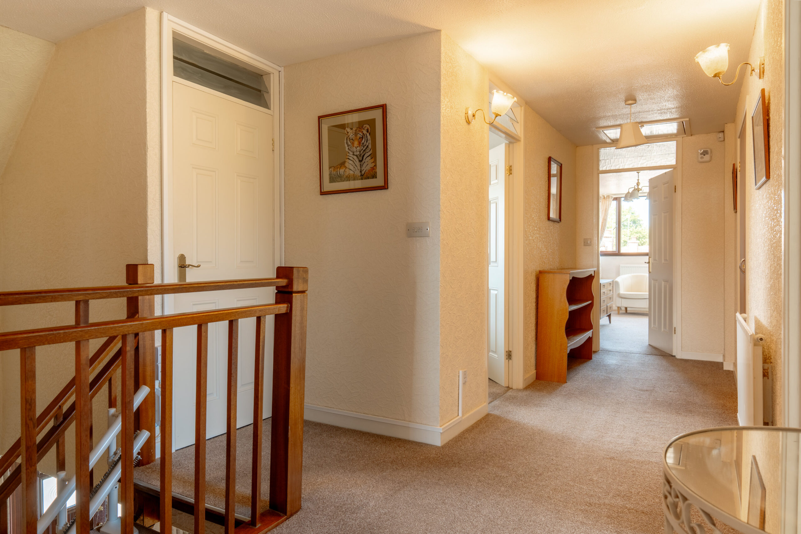 A well-lit hallway in a house with beige walls, carpeted floor, wooden stair railing, several doors, a framed tiger picture on the wall, and a small glass table in the corner. Light streams in from a distant window.