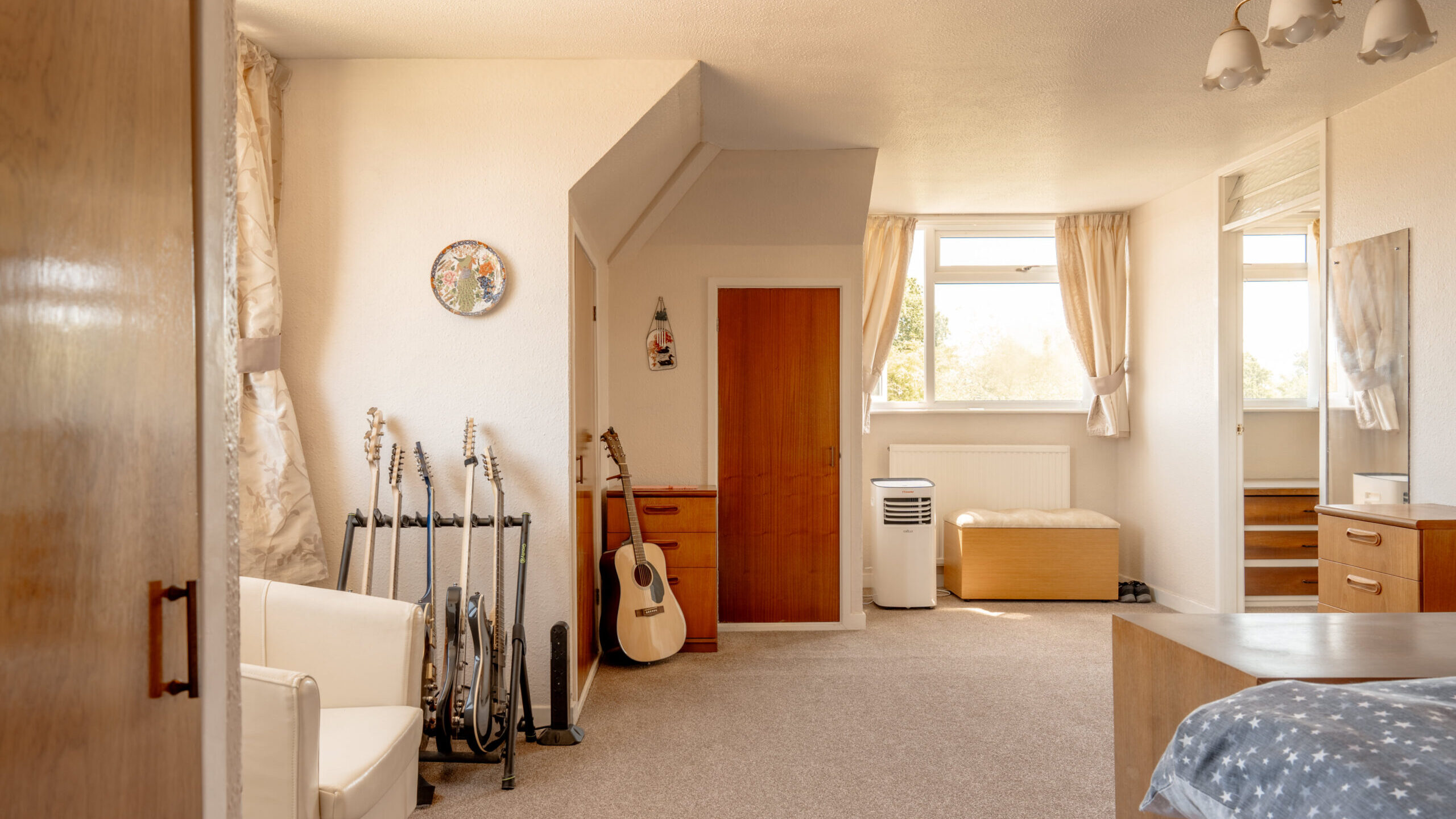 A bright bedroom with beige carpet, a bed with blue bedding, a white armchair, several guitars on stands, a wooden door, and large windows with cream curtains letting in natural light.