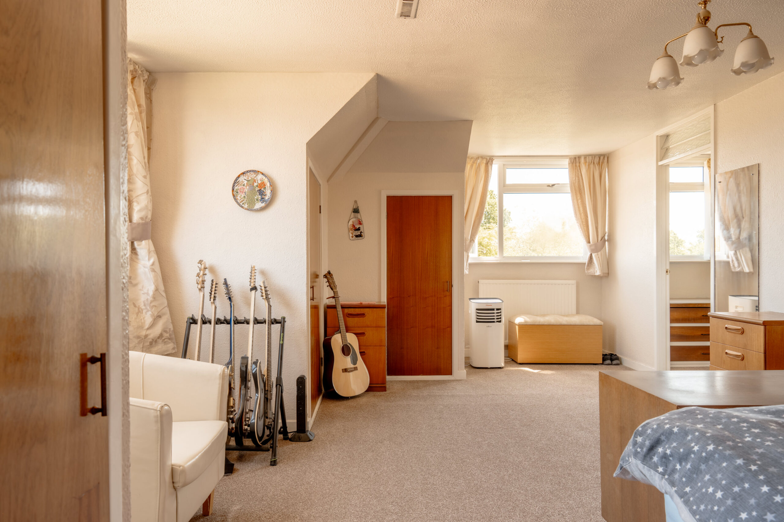 A bright bedroom with beige carpet, a bed with blue bedding, a white armchair, several guitars on stands, a wooden door, and large windows with cream curtains letting in natural light.