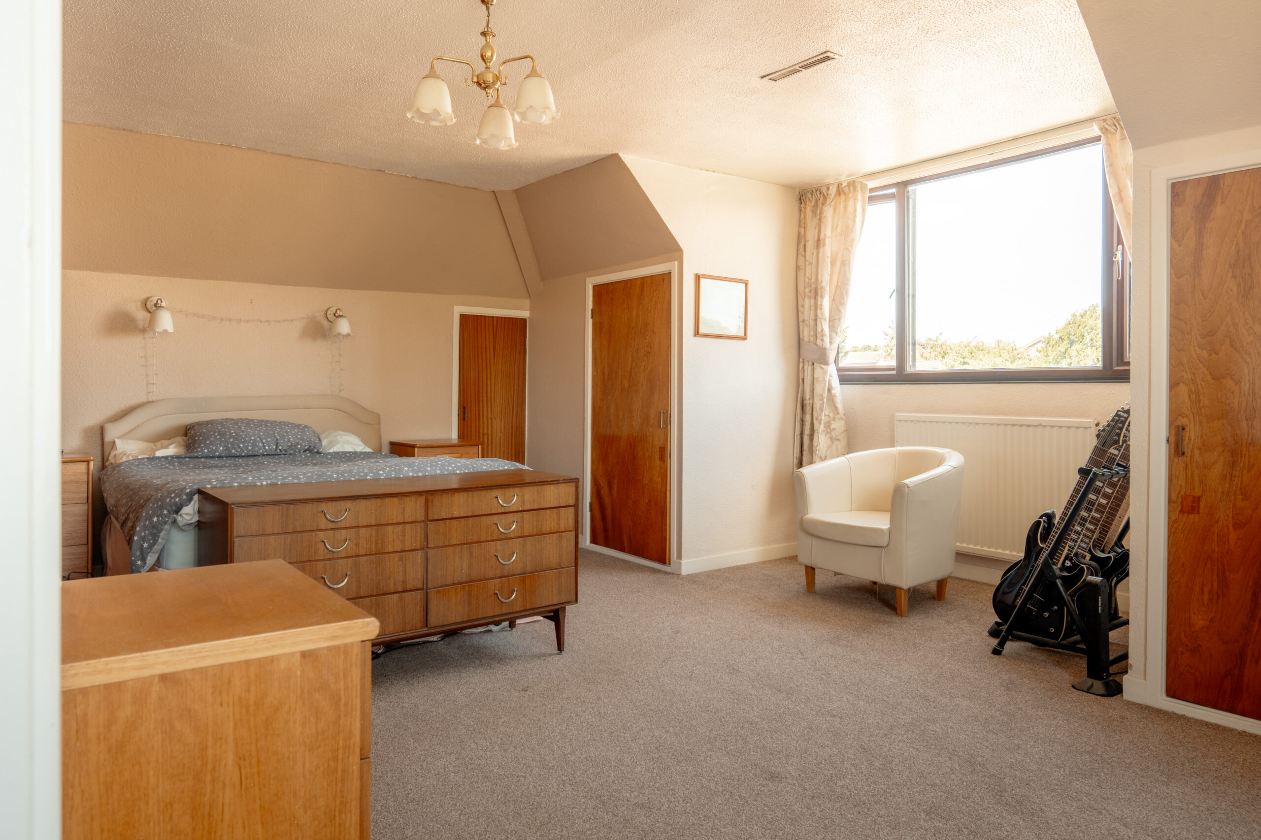 A spacious bedroom with a double bed, wooden furniture, a white armchair near a window, and two electric guitars with a stand in the corner. The room has beige walls and carpeted flooring.