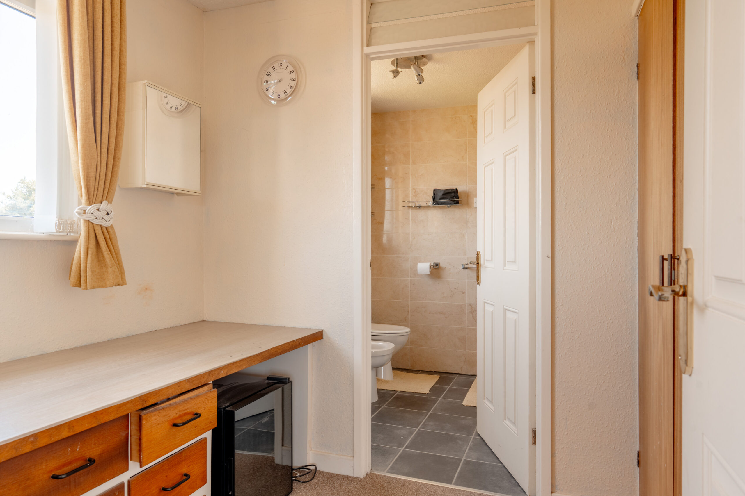A small room with a wooden desk and drawers, a mini fridge underneath, cream-colored walls, a window with gold curtains, a wall clock, and an open door leading to a beige-tiled bathroom.
