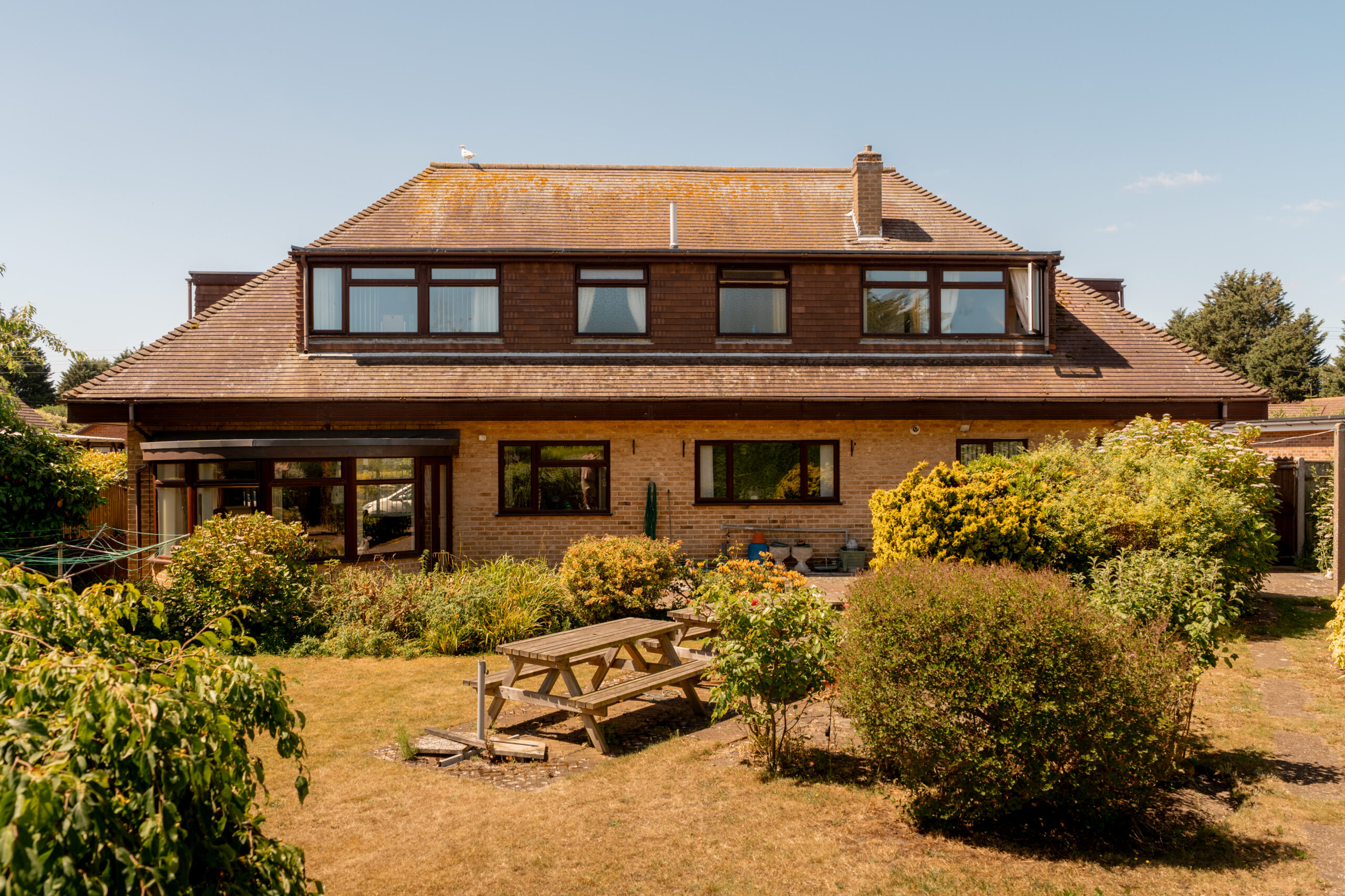 A large, two-story house with a brown roof and brick exterior, surrounded by a lush garden with green bushes and a wooden picnic table on a dry, yellowish lawn under a clear blue sky.