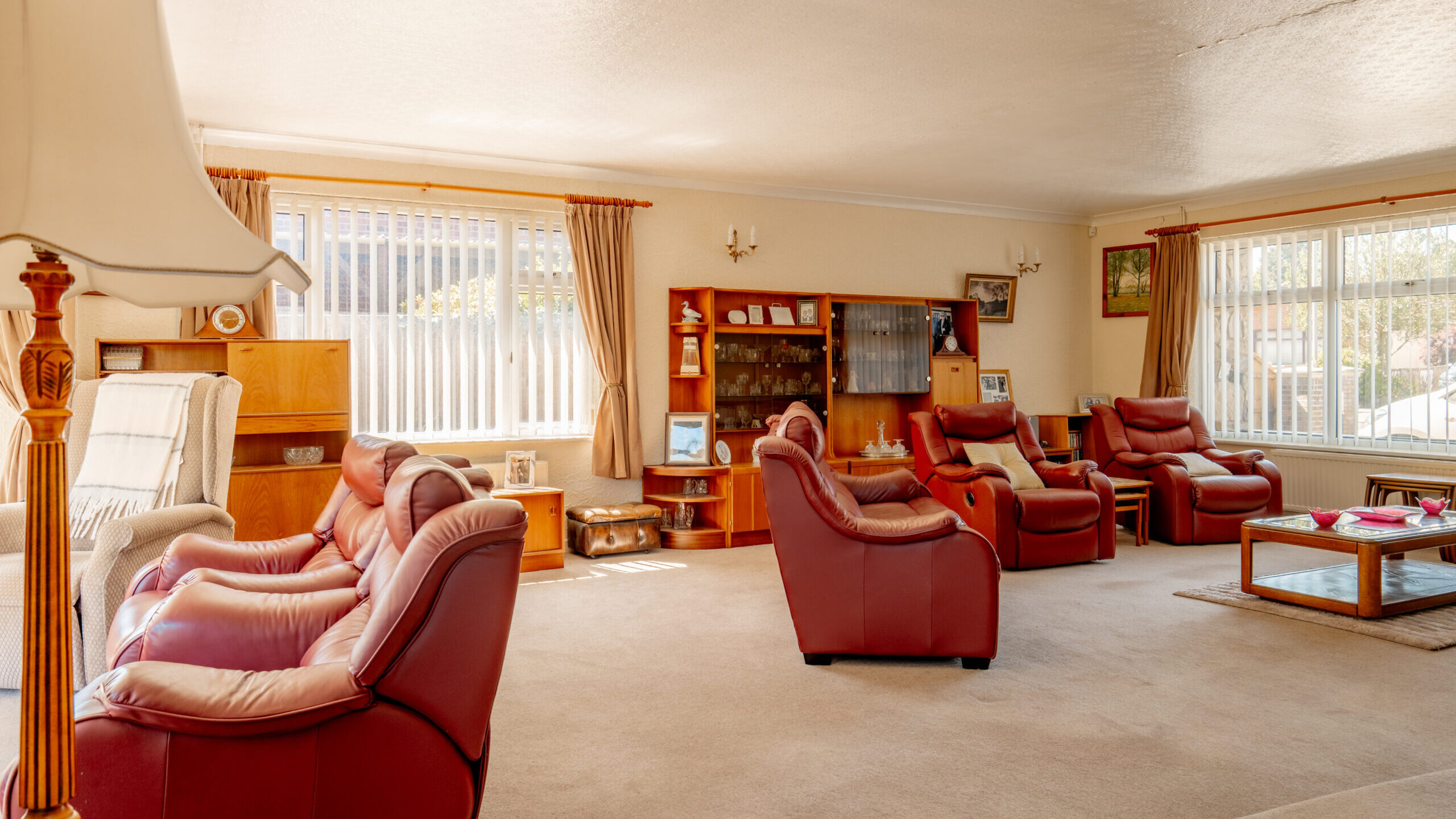 Spacious living room with red leather armchairs, wooden furniture, large windows with beige curtains, and a glass coffee table on a light carpet. Sunlight streams in, creating a warm and inviting atmosphere.