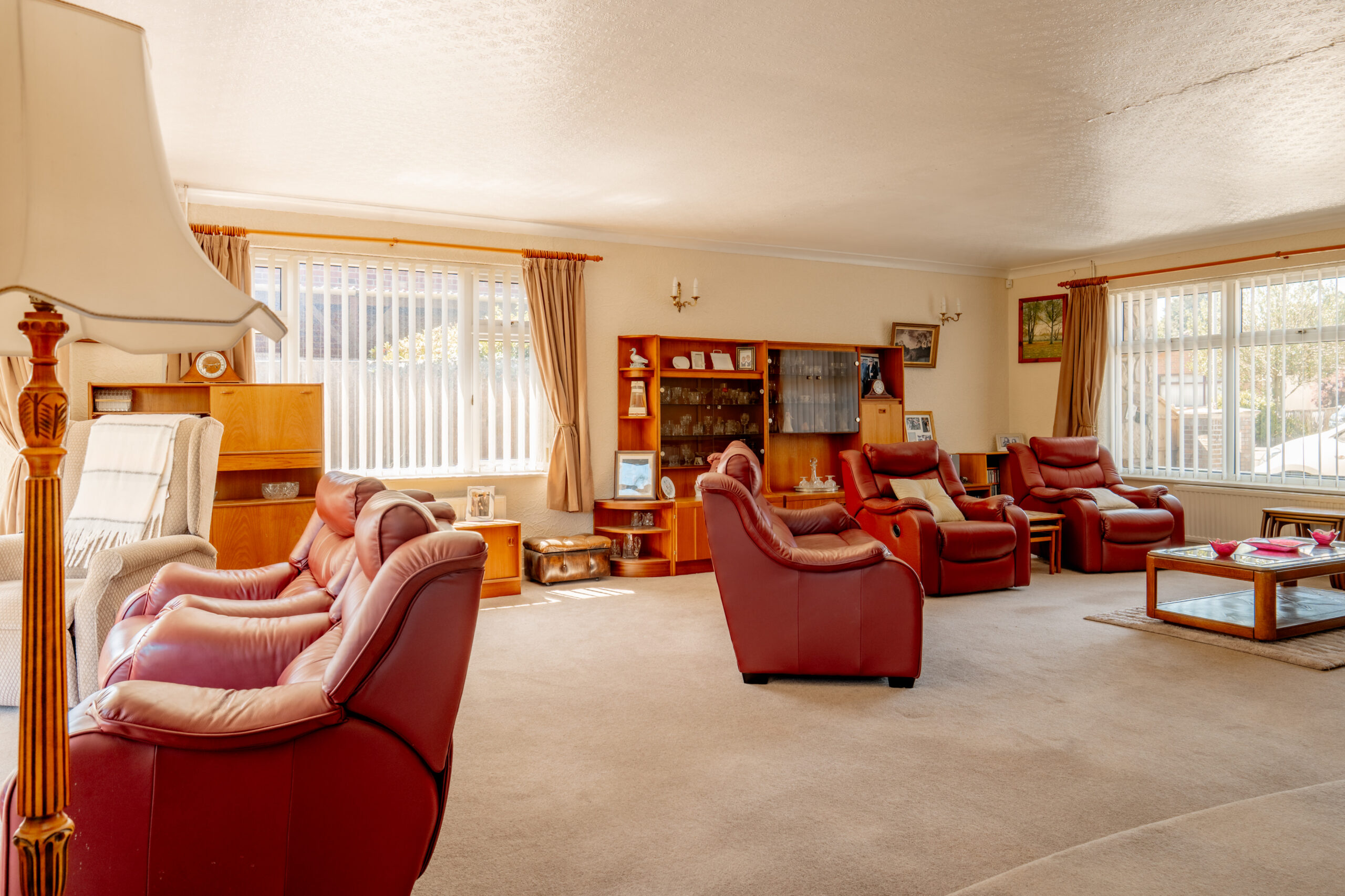 Spacious living room with red leather armchairs, wooden furniture, large windows with beige curtains, and a glass coffee table on a light carpet. Sunlight streams in, creating a warm and inviting atmosphere.