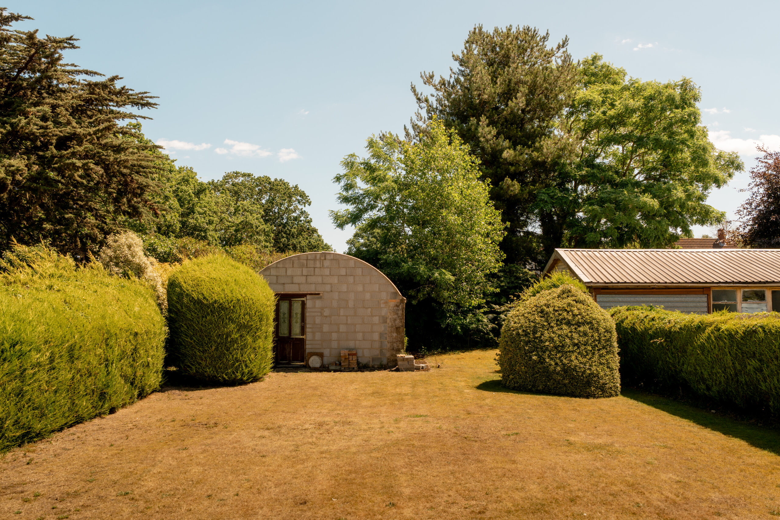 A small, arched concrete shed sits in a sunny backyard, surrounded by neatly trimmed bushes and large green trees. To the right, part of a house with a metal roof is visible. The lawn is slightly dry and patchy.