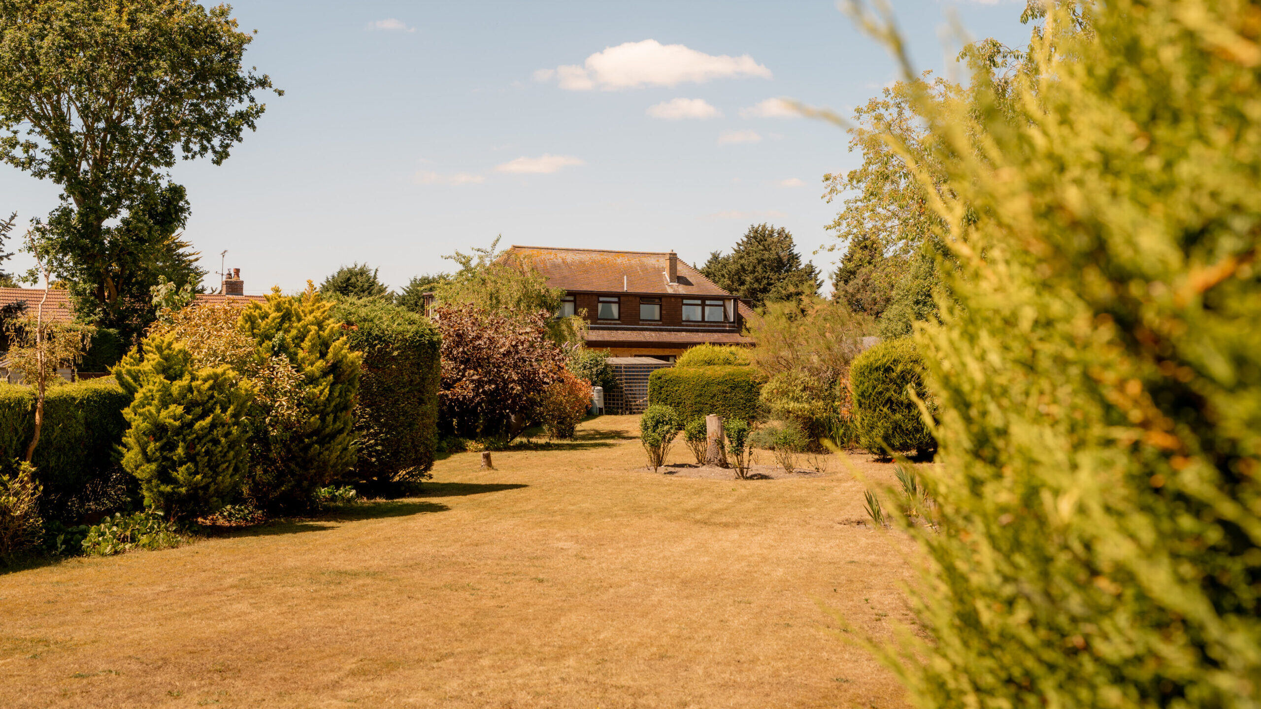 A large brown house with a sloped roof sits at the end of a spacious, sunlit garden with trimmed hedges and trees on a clear day with blue sky and scattered clouds.