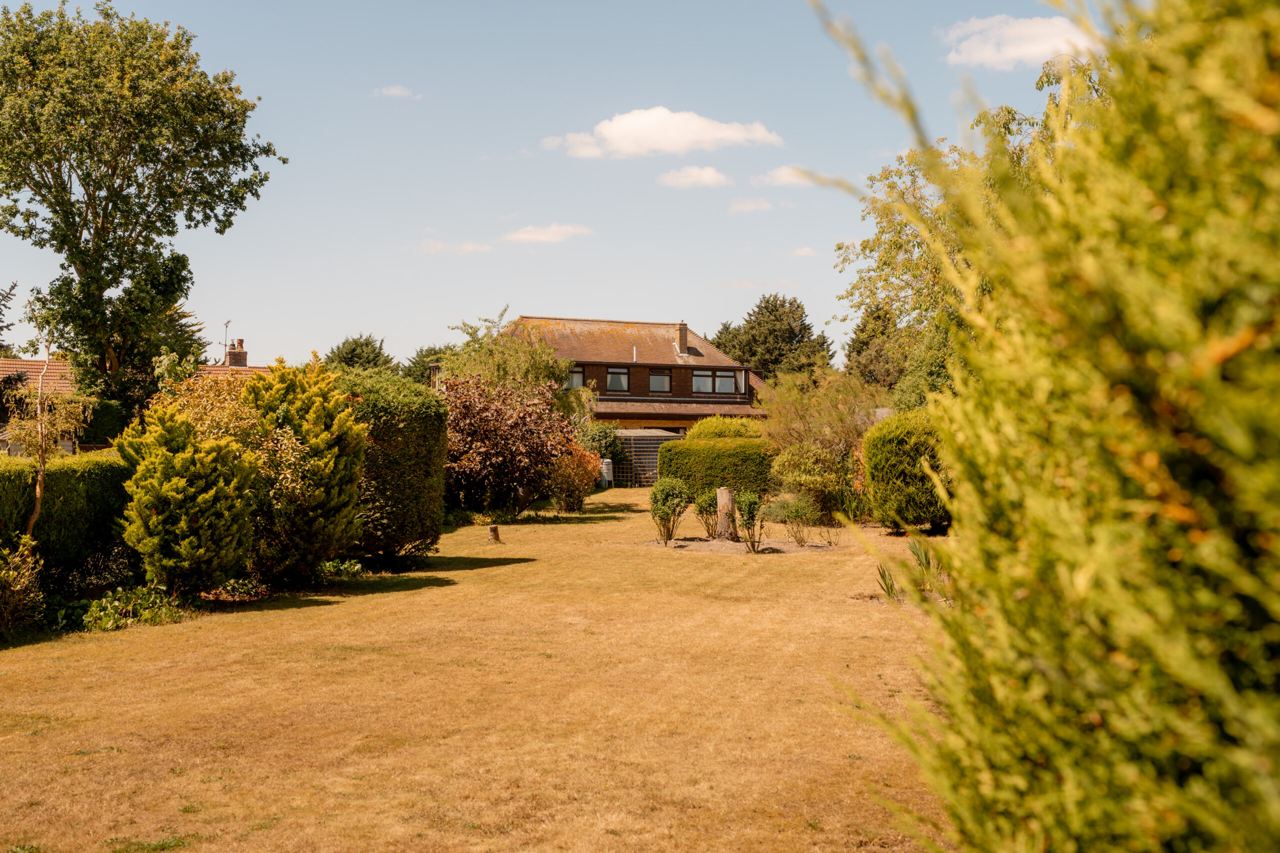 A large brown house with a sloped roof sits at the end of a spacious, sunlit garden with trimmed hedges and trees on a clear day with blue sky and scattered clouds.