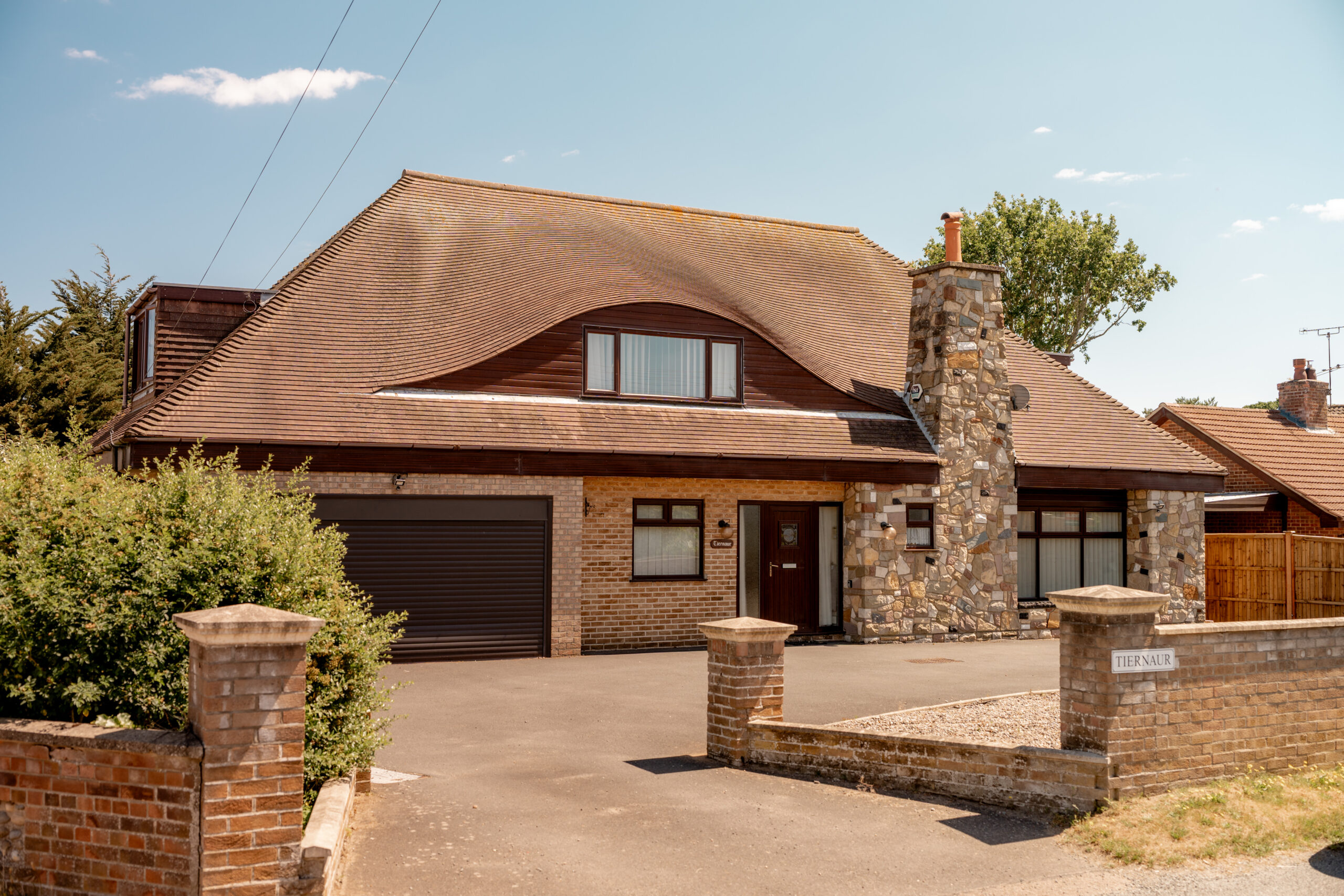 A modern two-story house with a unique curved roof, stone chimney, and large garage, set behind a brick gate on a sunny day. There is greenery and trees around the property.