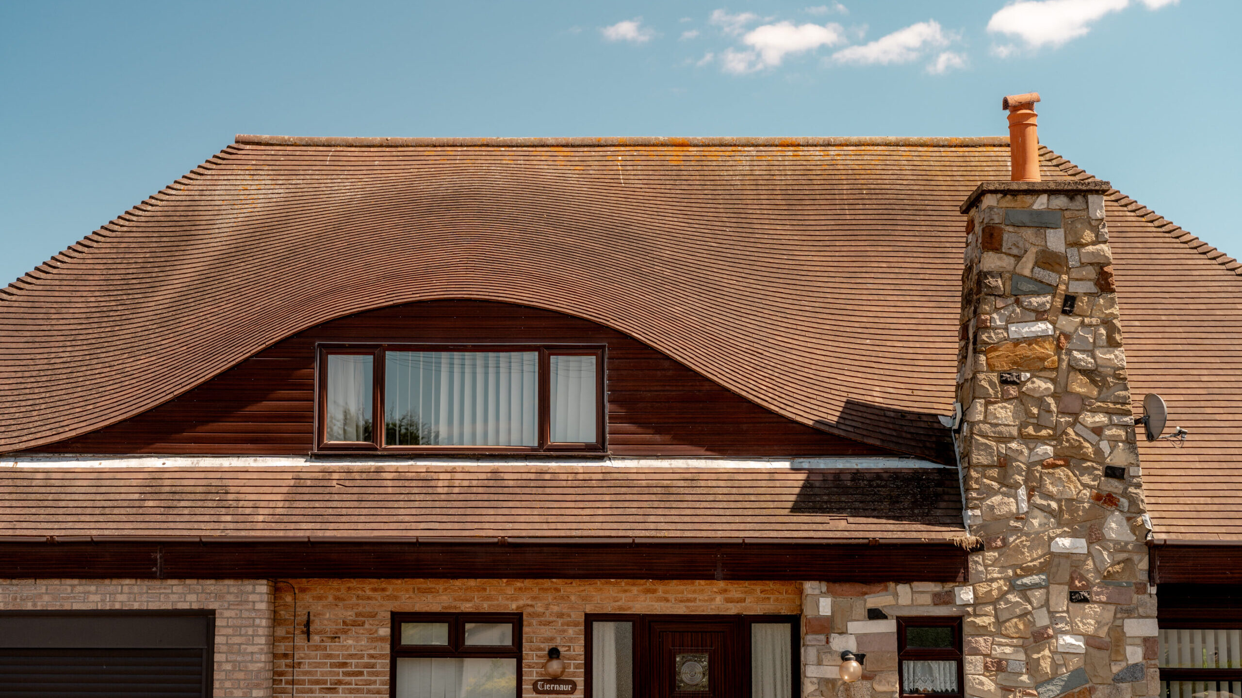 A house with a unique wavy tiled roof, a large stone chimney, brown wooden accents, and rectangular windows under a clear blue sky with a few small clouds.