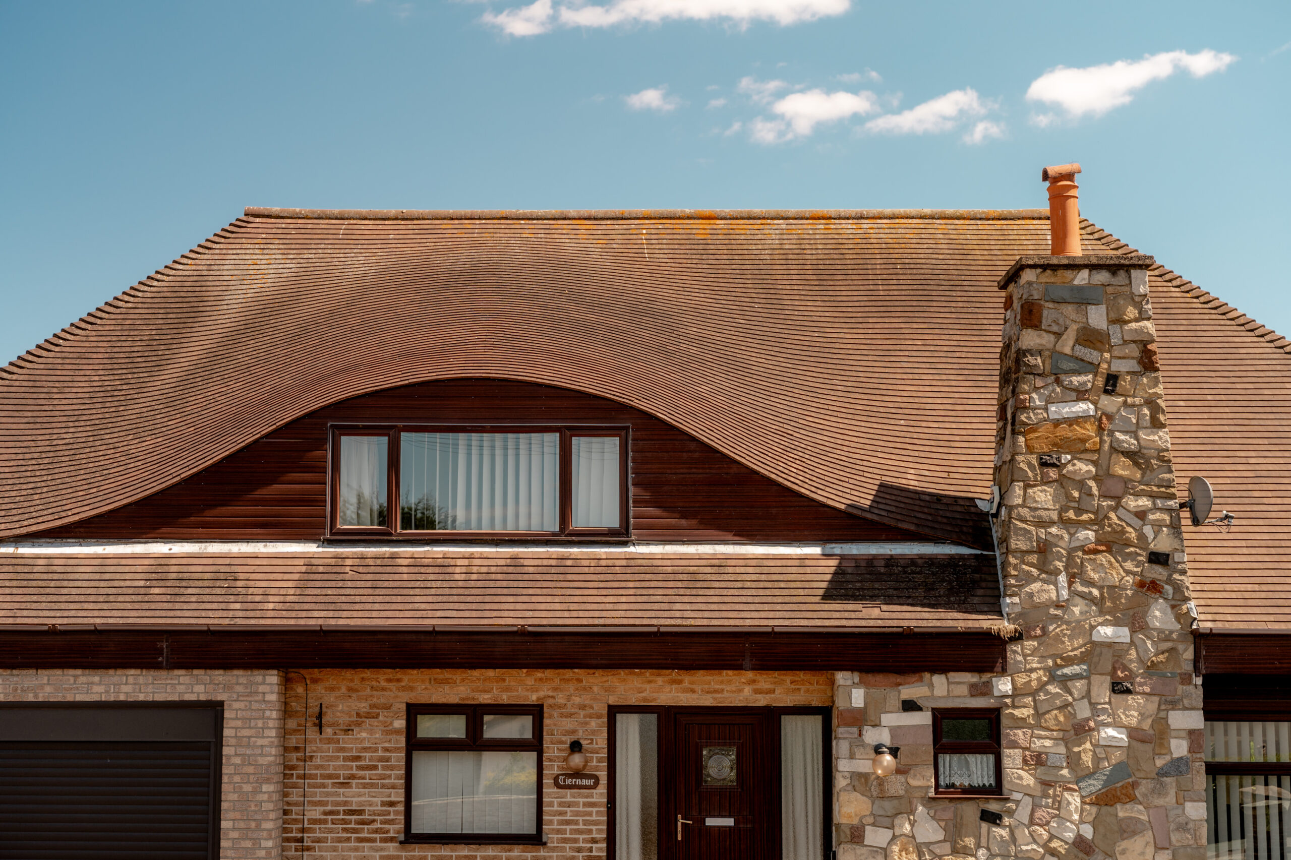 A house with a unique wavy tiled roof, a large stone chimney, brown wooden accents, and rectangular windows under a clear blue sky with a few small clouds.