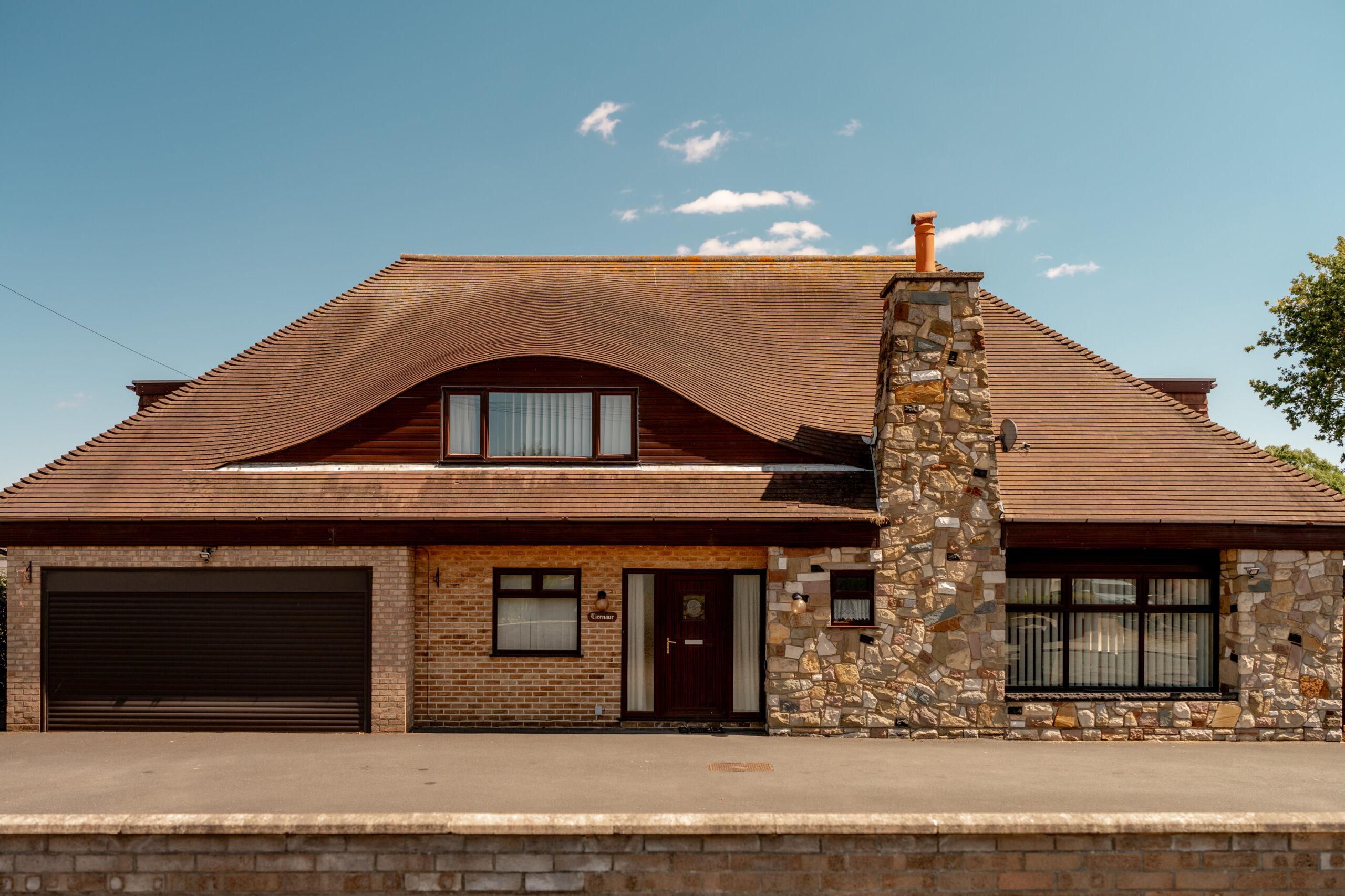 A modern house with a wavy, tiled roof, stone chimney, brick and stone facade, large windows, and a dark brown double garage door, set against a clear blue sky.