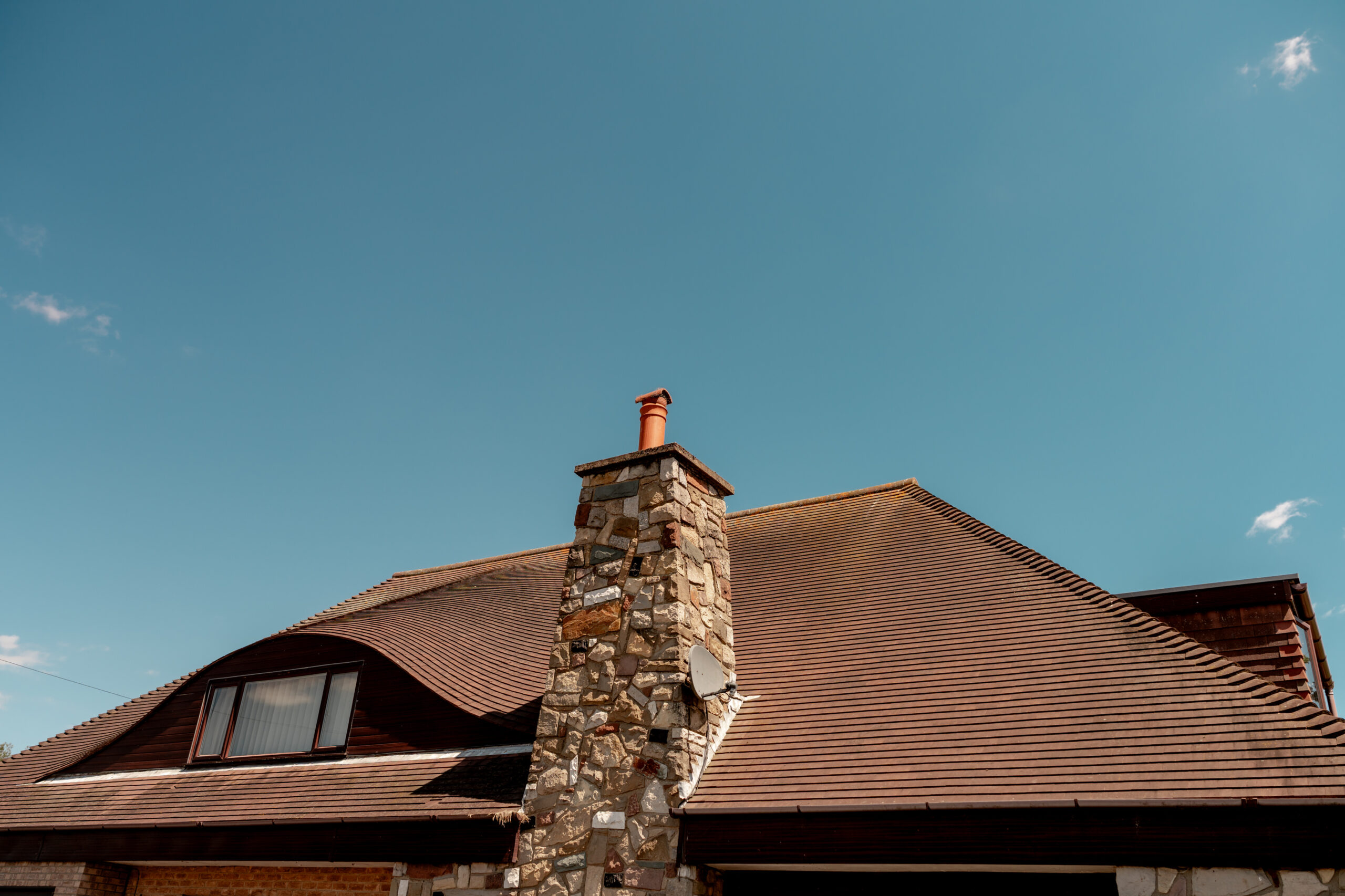 A stone chimney rises above a sloped, brown-tiled roof of a house with a large window, set against a clear blue sky.