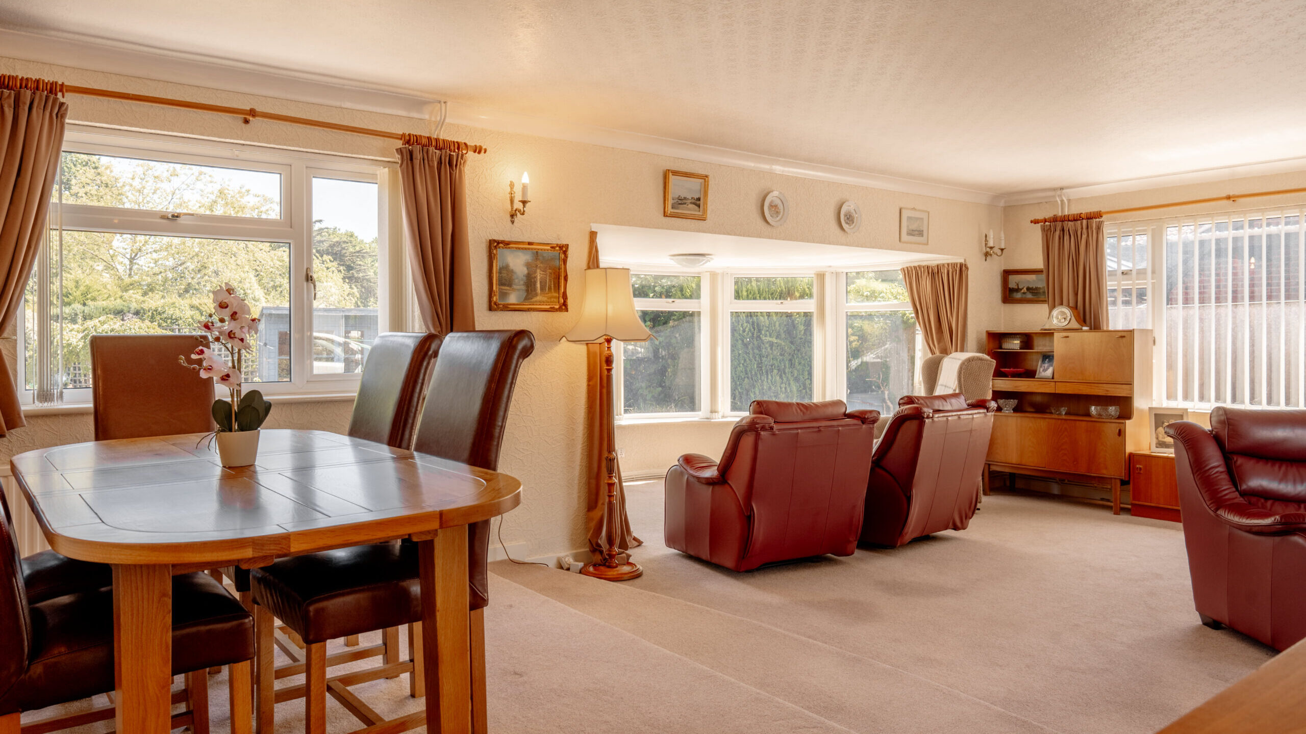 Spacious living and dining room with large windows, brown leather chairs, a wooden dining table with chairs, a lamp, and classic wooden furniture. Neutral carpet and light-colored walls create a warm, inviting atmosphere.