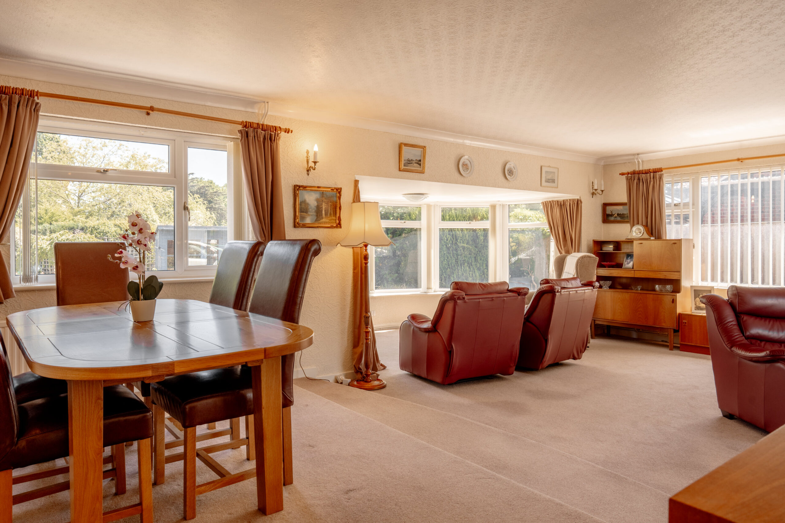 Spacious living and dining room with large windows, brown leather chairs, a wooden dining table with chairs, a lamp, and classic wooden furniture. Neutral carpet and light-colored walls create a warm, inviting atmosphere.