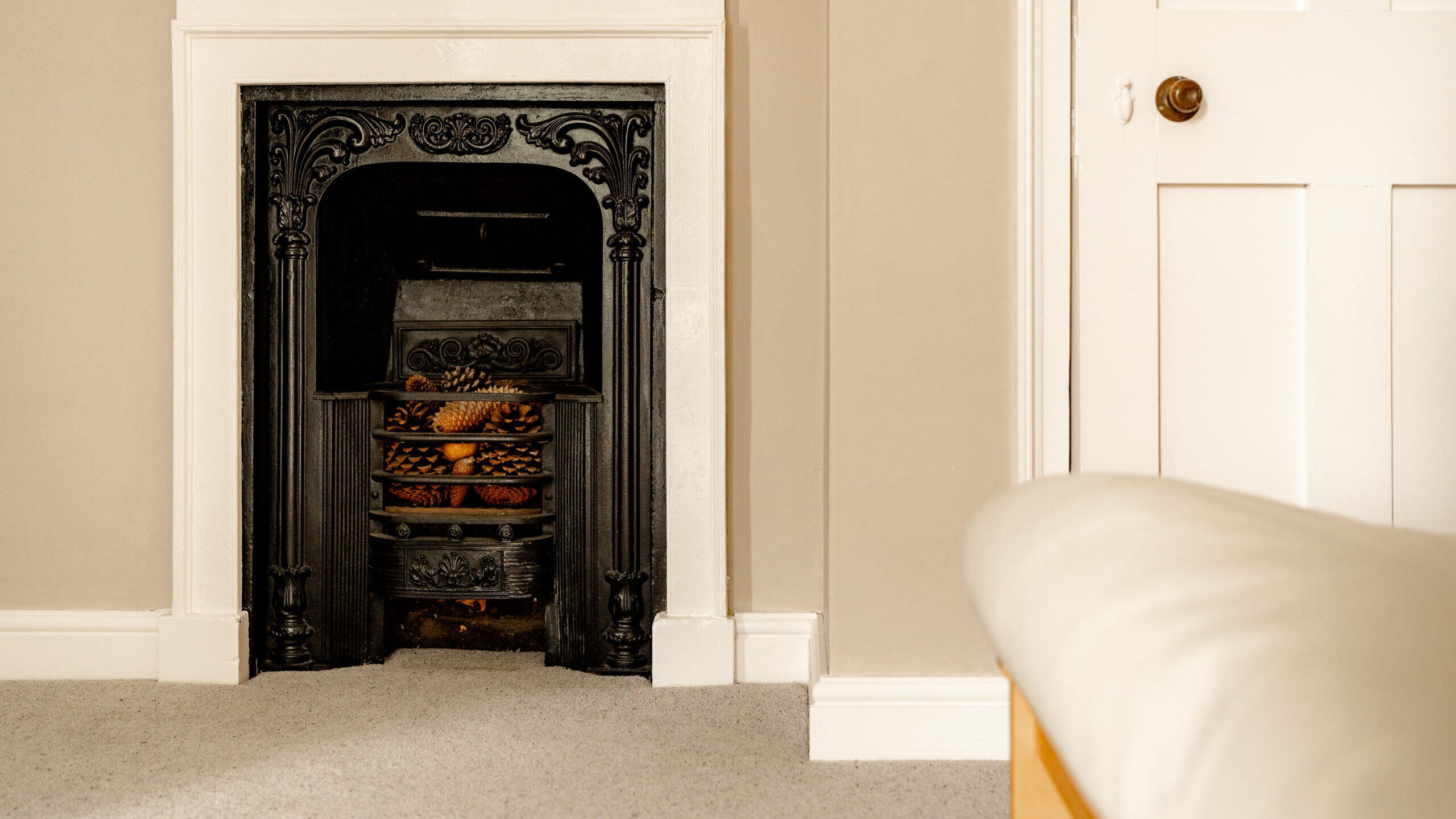 A black ornate fireplace with pinecones inside sits against a beige wall, next to a closed white door and a bed with a white comforter on a carpeted floor.